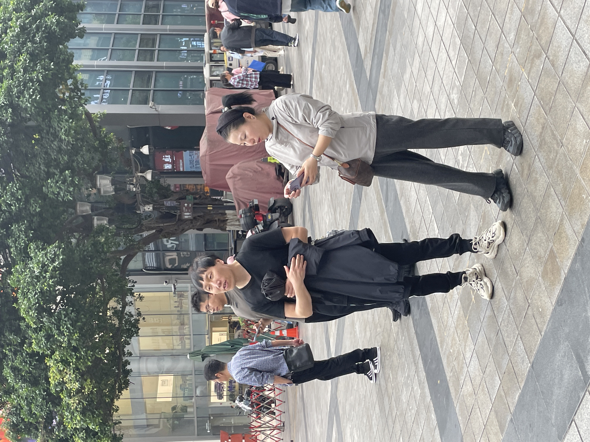 A group of pedestrians are walking in an outdoor urban environment. There is a woman wearing a light grey jacket and dark grey pants, holding a phone in her hand, and wearing a brown crossbody bag. There is a man wearing a black t-shirt, black pants, and carrying a black coat. A third person can be seen behind him. In front, there is a man wearing a blue and white checkered shirt with black pants and black shoes. The background includes a glass building, trees, and a paved ground in a diamond pattern. A partial view of retail stands can be seen. Based on the architecture and street design, this appears to be the Yuzhong District of China.
