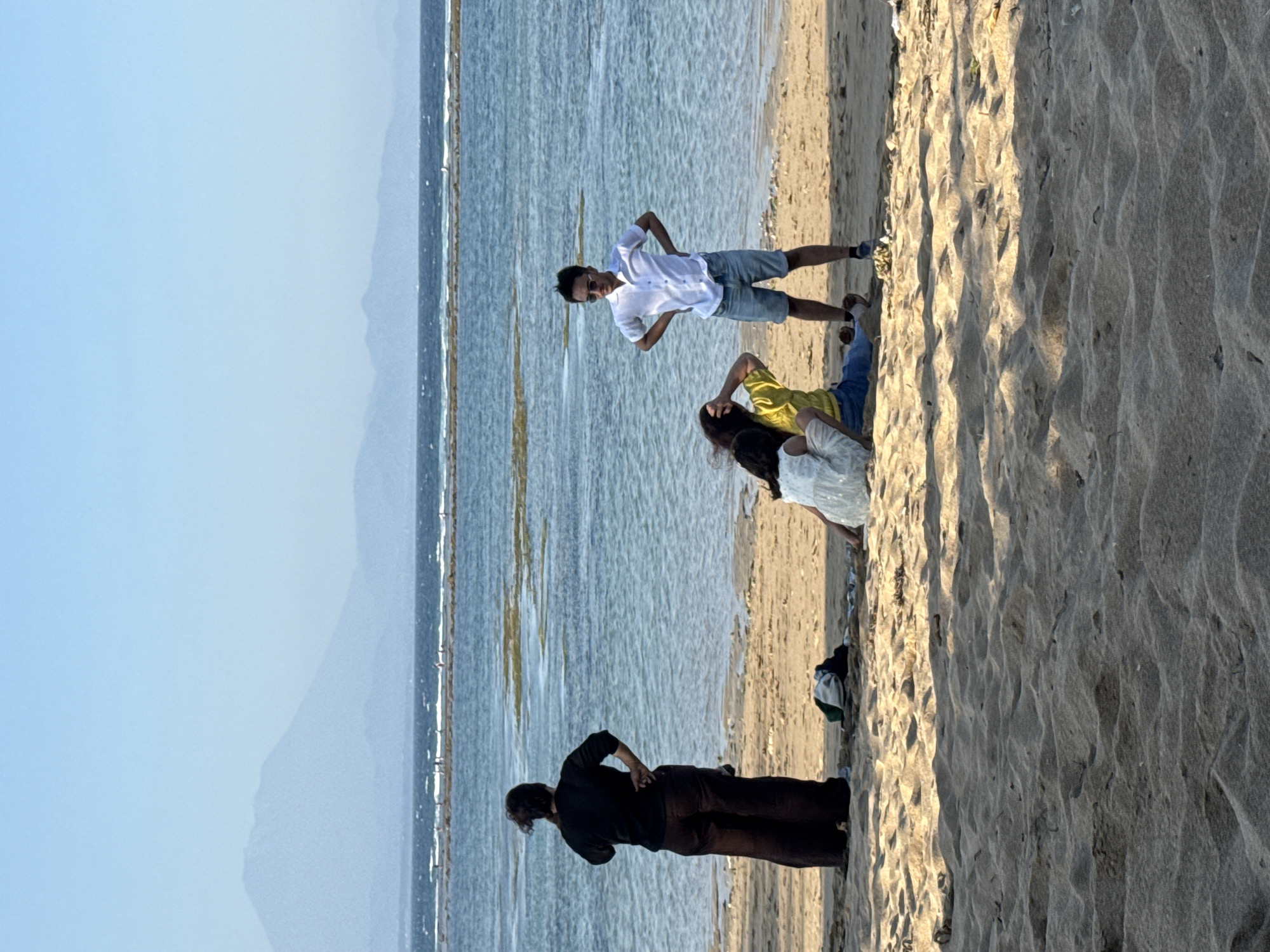 In Denpasar, Indonesia, on a sandy beach, four individuals are present near the water's edge under a clear blue sky. To the left, a person stands with their back to the camera, wearing a dark top and wide-leg pants. In the center foreground, two individuals are seated on the sand; one wears a white garment and the other a yellow top and blue pants. To their right, a man stands facing them, wearing a white button-down shirt and denim shorts, with his hands on his hips. The ocean stretches out behind them, with a distant mountain range visible on the horizon. Several small boats or structures are visible in the water, along with shallow areas that appear to have vegetation. Bags and clothing are scattered on the sand in the foreground.
