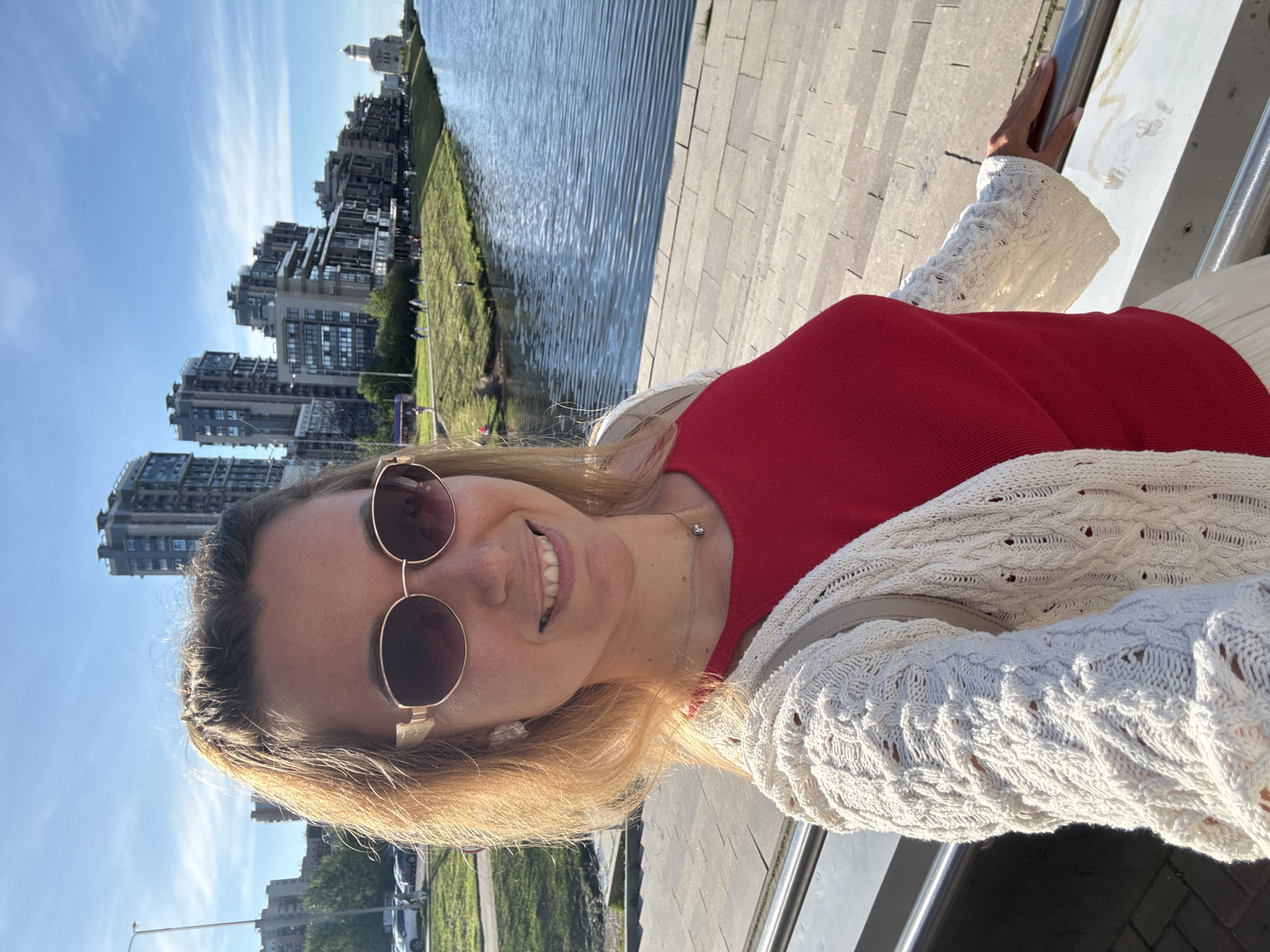An outdoor, waist-up, selfie-style, medium shot shows a blonde woman with light skin, wearing sunglasses with rose-gold rims and a cream-colored knit cardigan over a red top. She is smiling and facing the camera, with her right hand resting on a metal railing. The background shows a body of water, a grassy bank with people walking, and a line of modern, multi-story buildings. The sky is blue with scattered clouds. The scene appears to be a waterfront promenade in a city.
