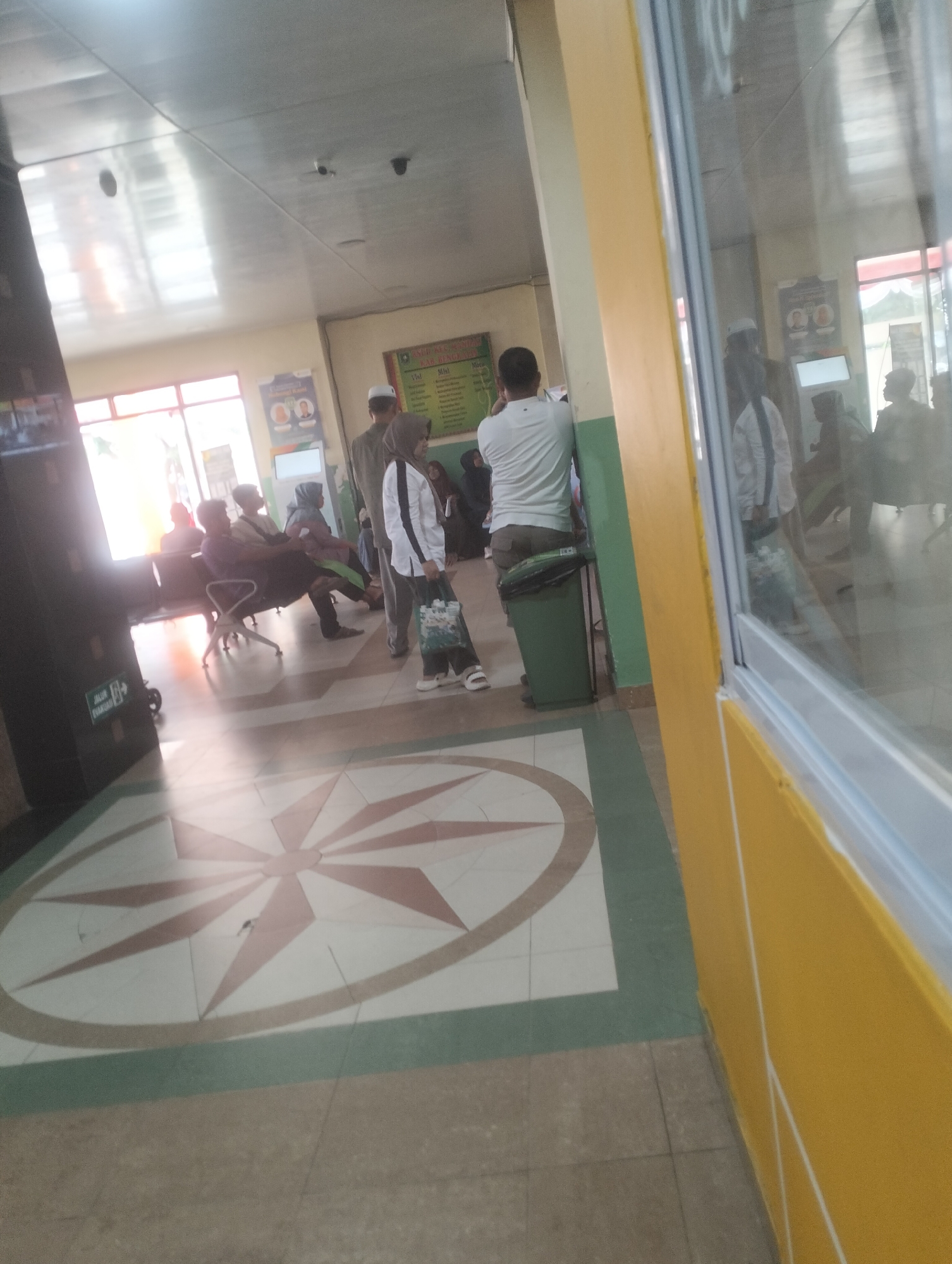 The image depicts an indoor waiting area, possibly a clinic or a public service office, in Air Jamban, Indonesia. The scene is illuminated by overhead fluorescent lighting, casting a bright glow on the tiled ceiling. In the foreground, a distinctive floor mosaic features a starburst pattern with alternating pink and white triangular sections, bordered by a circular band. To the right, a prominent yellow wall runs parallel to a large glass pane, reflecting the interior space and the people within.
Several individuals are present in the waiting area. On the left, a group of people are seated on dark-colored benches. Among them, a person wearing a blue shirt is visible. Further to the right, a person in a light-colored shirt and dark pants stands facing away from the camera, holding a patterned tote bag. Adjacent to this person is a green trash can. In the mid-ground, a woman in a hijab and a black and white striped shirt is walking away from the viewer. Other individuals, some also wearing hijabs, are partially visible in the background, some seated and others standing.
On the wall, a poster with Indonesian text and imagery of two figures is displayed. Another poster, with green and white text, is also visible. A dark object, possibly a cabinet or kiosk, is positioned on the far left of the frame, featuring a green sign with white text that reads "JALUR EVAKUASI" (Evacuation Route). The overall environment suggests a functional, public space designed for waiting and service.