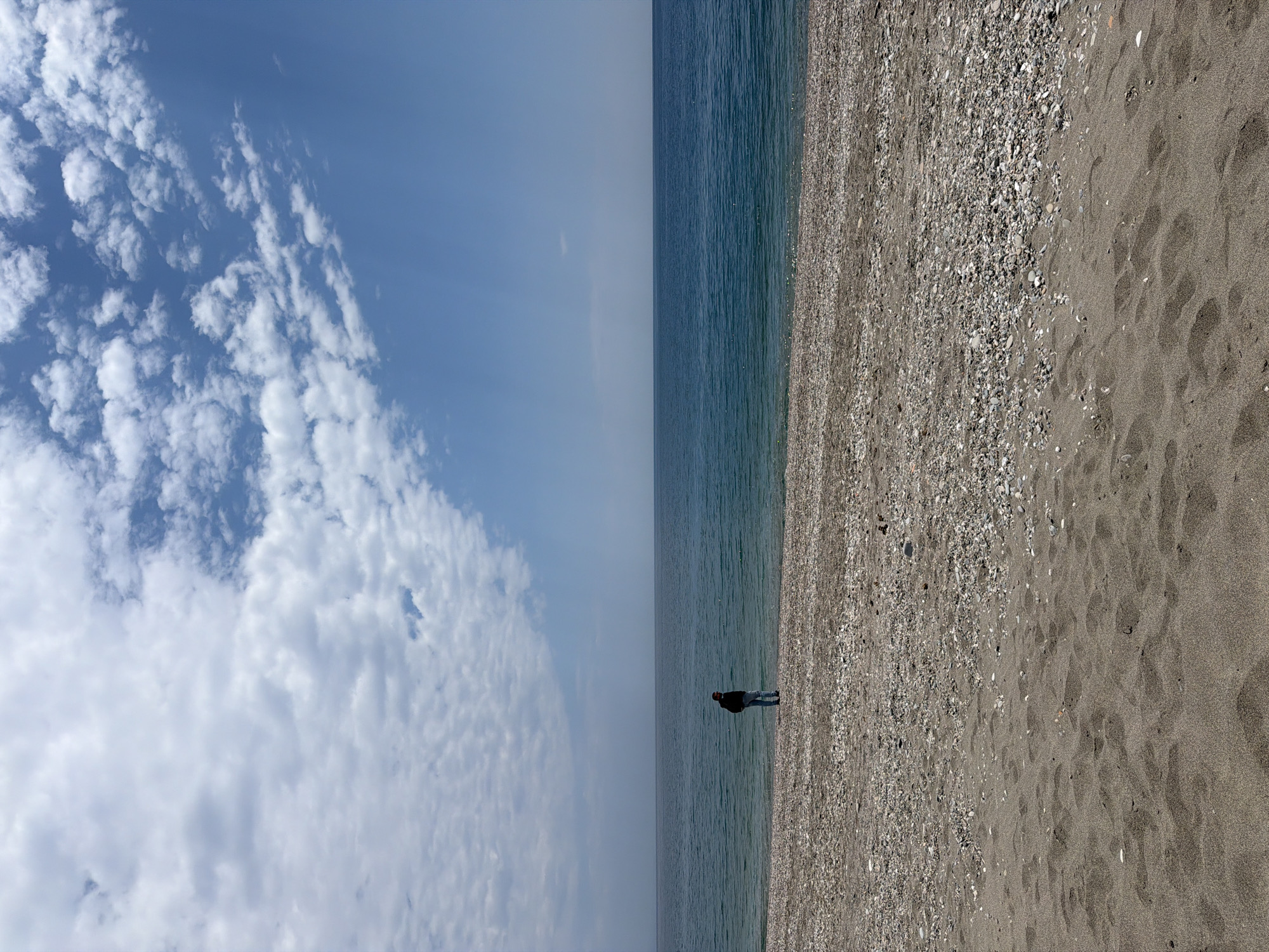 A horizontally oriented photograph shows a person standing on a beach next to the sea. The scene is possibly in Chianchitta, Italy, although this is not confirmed by visual evidence. The left half of the image shows a bright blue sky partially covered with fluffy, white cumulus clouds. The sky fades into a hazy, light blue near the horizon. The right half of the image depicts a beach with darker, greyish-brown sand transitioning to a lighter, more pebbly sand near the water's edge. A single person, appearing to be wearing dark clothing, is standing on the beach near the water. The ocean water is a dark, slightly muted blue-green. The camera angle is slightly elevated and directed downwards. The image is sharp and well-lit.