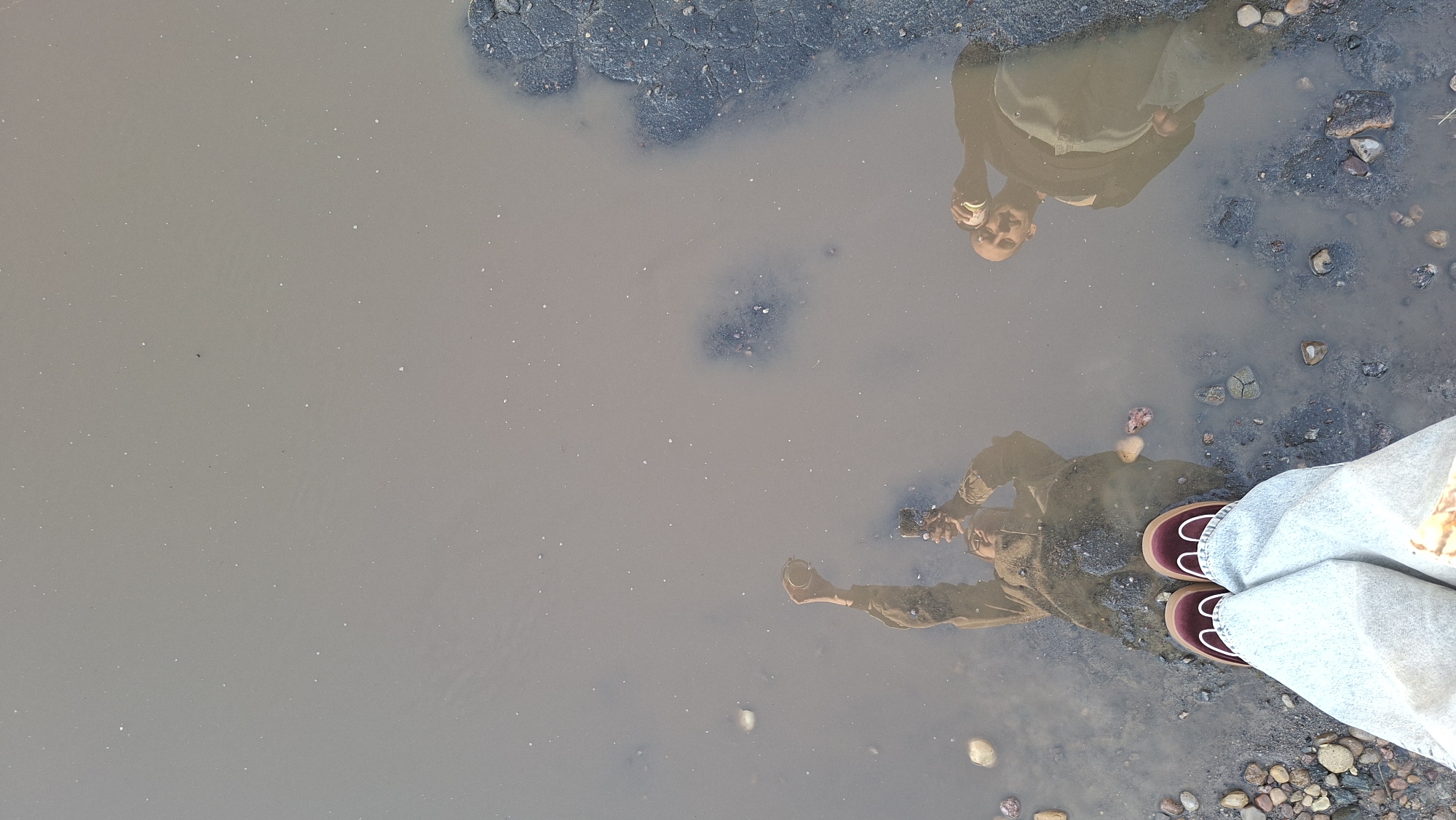 The image is a reflection in a puddle. Two individuals are visible in the reflection. One person, on the left, is holding a phone up, appearing to take a photo. This person is wearing a tan jacket and jeans, and dark shoes with white trim. The second person, on the right, is holding a can and looking towards the camera. This person is wearing a similar tan jacket and appears to be bald. The puddle is situated on a surface of gravel and broken asphalt, consistent with a road or path. The environment suggests an outdoor setting, potentially in or near Забалоцкі сельскі Савет, Belarus, based on the provided location context. The water is murky and reflects the sky and the surrounding environment.