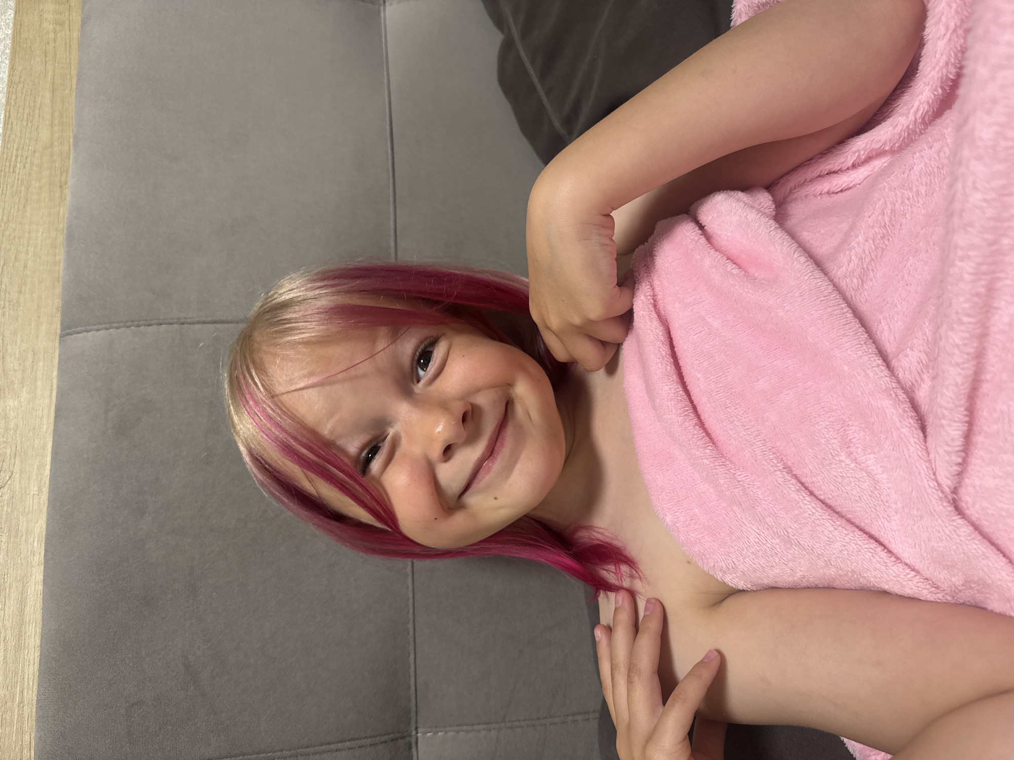 A young child with light blonde hair and pink streaks lies on a gray couch, wrapped in a pink fuzzy blanket. The child is smiling and looking at the camera. Part of a darker gray object is visible in the upper right corner of the image. The location is unknown.