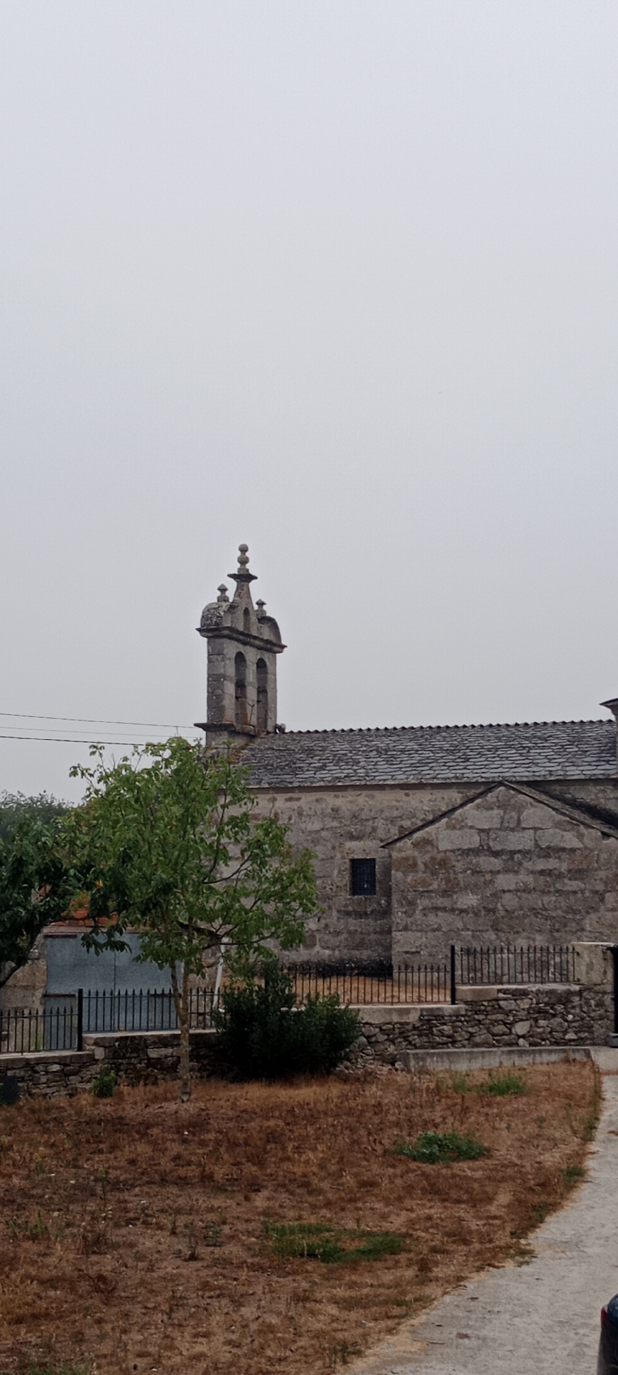 The image depicts a cloudy day in Adai, Spain, with a stone building that appears to be a church as the central subject. The church has a distinctive bell tower with two arched openings and a decorative finial. The roof of the church is covered with dark, irregular stone tiles, and the walls are constructed from rough-hewn grey stones. A small, dark rectangular window is visible on the side of the building. In the foreground, a small green tree with sparse foliage stands to the left, and a patch of dry, brown grass covers the ground. A black metal fence with vertical bars runs along the front of the property, separating it from the dry grassy area. A stone wall with a concrete cap runs below the fence. A paved path or road is visible on the right side of the image. The overall impression is a rural or village setting.