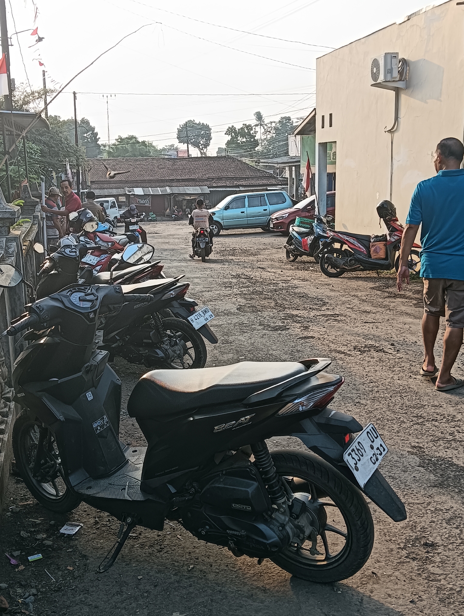 The image depicts a street scene in Cluwak, Indonesia, likely a commercial or residential area given the presence of multiple parked motorcycles and a building. In the foreground, a black scooter is parked facing left, with its license plate clearly visible. The license plate reads "3369 OU 02-31". Further into the scene, several other motorcycles are parked on a gravelly surface. A man wearing a blue t-shirt and tan shorts stands facing away from the camera, walking towards the right side of the image. Behind him, a white building with an air conditioning unit mounted on its exterior wall is visible. The background shows a row of buildings with tiled roofs and some trees. The sky is bright, suggesting daylight. The overall impression is a typical street scene in a town or city in Indonesia, with a focus on the parked motorcycles and the presence of a pedestrian.