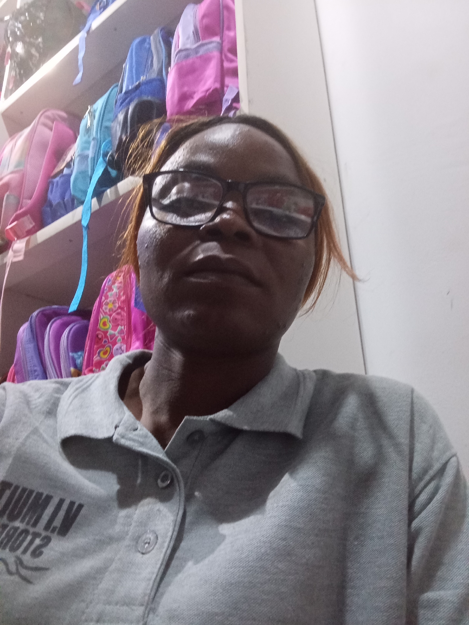 A young woman, wearing glasses and a grey polo shirt, poses for a selfie in front of shelves filled with backpacks. The background shows several brightly colored backpacks, suggesting a school or retail environment. The woman's hair is a light brown, and she has a warm complexion. The lighting is dim, with shadows casting across the scene, and the overall setting appears to be indoors. The image is taken from a close-up perspective, focusing on the woman's face and upper body. There is text visible on her shirt, but it is partially obscured.