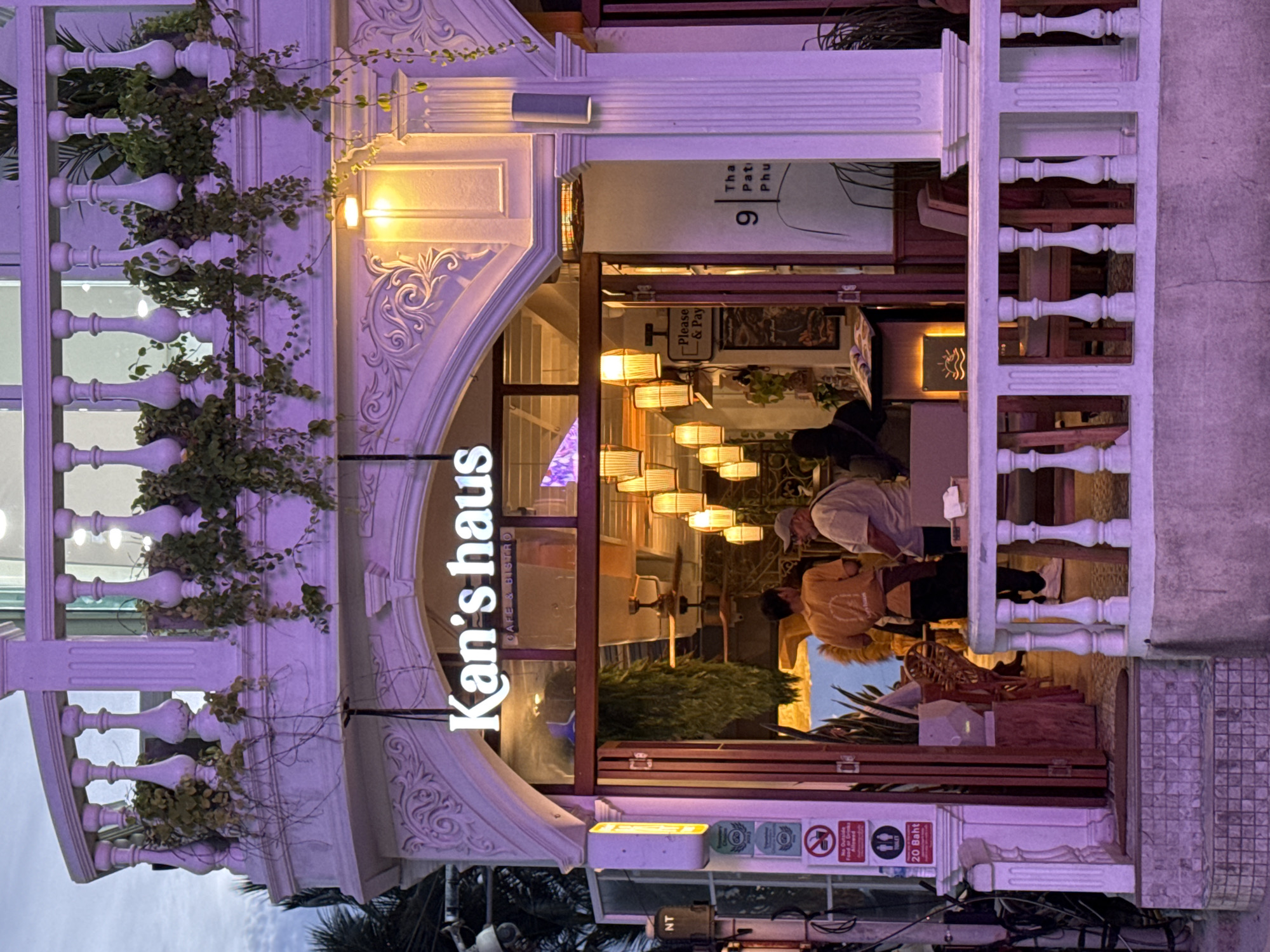 The image shows the exterior of a restaurant and cafe named "Kan's Haus" in Patong, Thailand. The building features a white, ornate facade with a balcony railing and decorative carvings. A large arched entrance reveals the interior, which is lit by numerous pendant lamps with bamboo shades. Inside, patrons can be seen dining and interacting. There are signs posted outside the entrance, including one with the text "Please & Pay" and another indicating "20 Baht" and a toilet symbol. The lighting suggests it is either dusk or evening, with the exterior illuminated by warm artificial lights and a purple hue from ambient lighting.