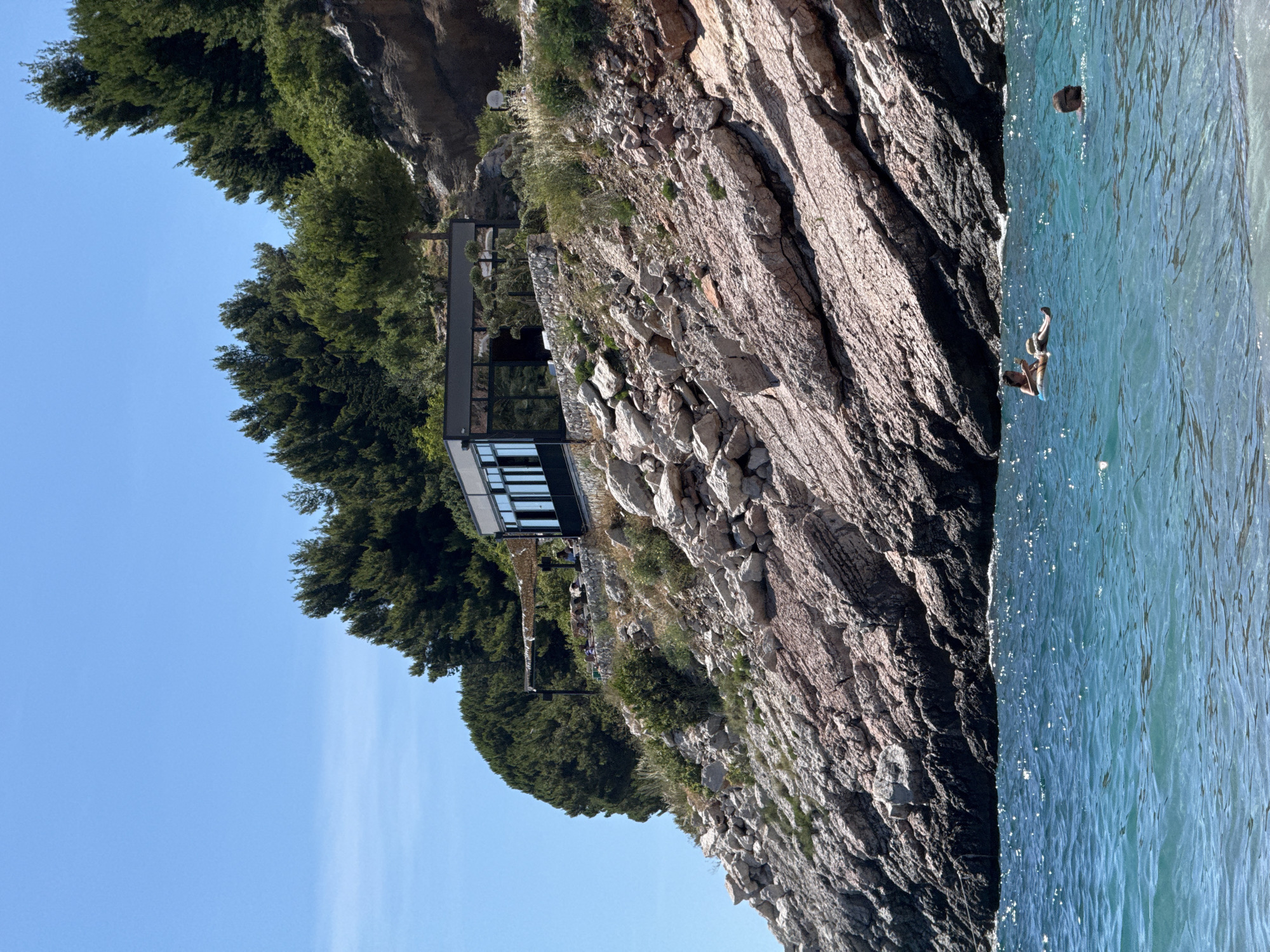 The image depicts a coastal scene in Kamenovo, Montenegro. In the foreground, the turquoise water of the Adriatic Sea shimmers under a clear blue sky. Two individuals are visible in the water: one person is relaxing on an inflatable float, while another is swimming.
Dominating the midground and background is a rocky, steep cliffside covered in dense green pine trees. Nestled on the cliff, amidst the rocks and vegetation, is a modern building with a dark-colored roof and large glass windows. A covered outdoor seating area is attached to the building, with several people seated there, suggesting it may be a restaurant or cafe. The rocky terrain is characterized by layers of reddish-brown and grey stone, with scattered vegetation growing in the crevices. The overall impression is a serene coastal landscape with a modern architectural element integrated into the natural environment.