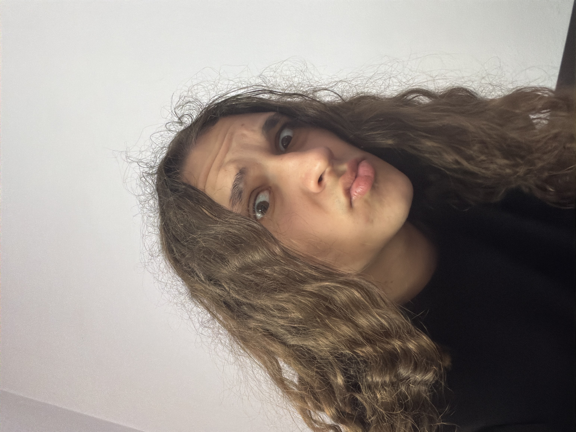 A close-up, tilted portrait features a young person with light skin, brown curly hair with frizz around the forehead, and a slight pout on their lips. Their eyes are wide and expressive, and their eyebrows are slightly furrowed. They are wearing a black garment. The background is a plain white wall. There is no indication of the location being Asenovgrad, Bulgaria.