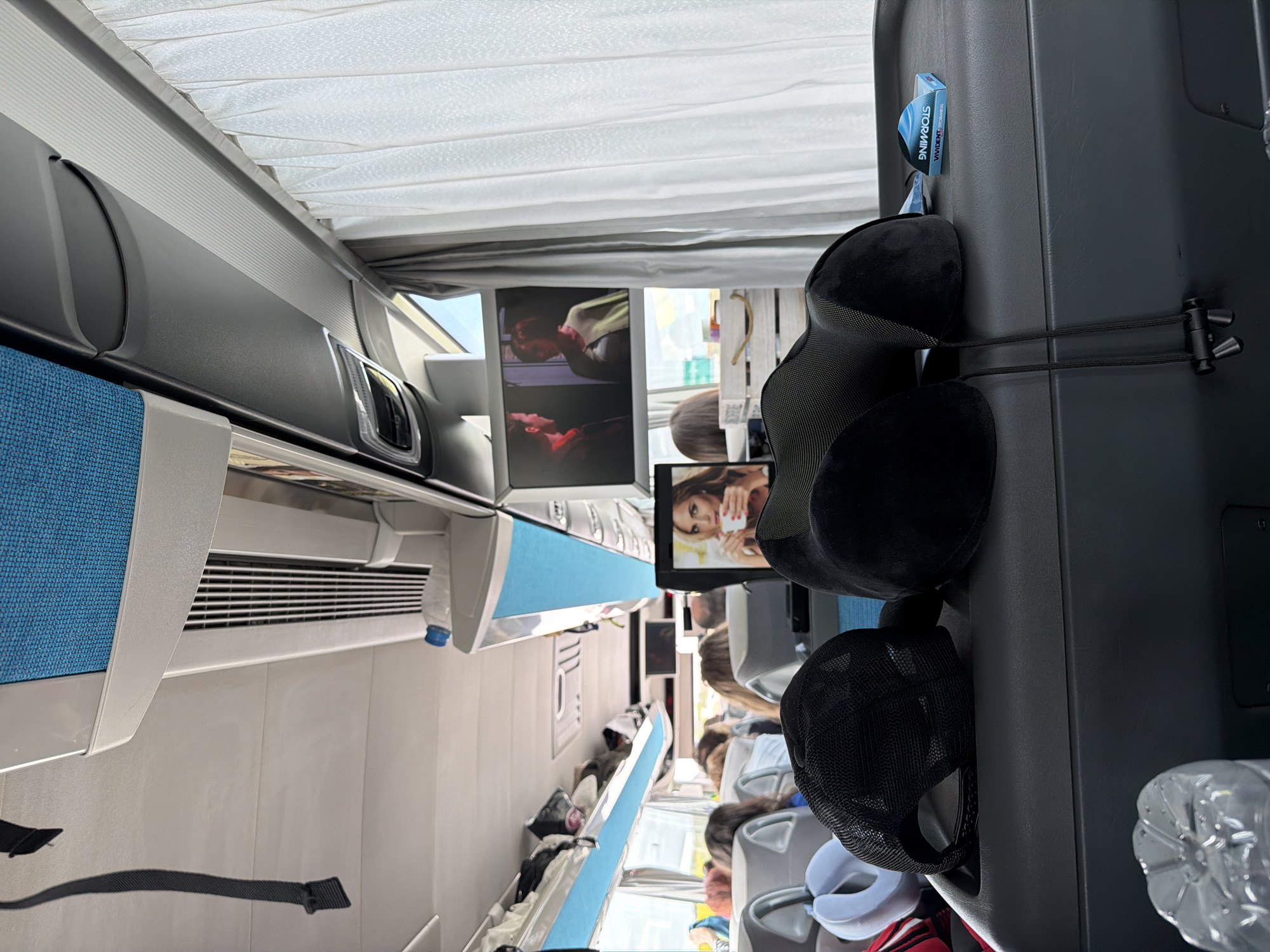 The image depicts the interior of a passenger vehicle, likely a long-distance bus or train, viewed from a seated passenger's perspective. The scene is oriented to show the aisle stretching into the distance towards the front or middle of the cabin, with rows of seats on either side.
In the immediate foreground, on the back of the seat directly in front of the viewer, a black memory foam travel pillow and a black mesh baseball cap are hanging. A small, partially obscured clear plastic water bottle is visible next to these items. Clipped to the seat back, above the travel pillow, is a small blue object with the text "VIVIDENT" and "STORMING" printed on it.
Further down the aisle, several individual entertainment monitors are visible on the backs of seats. These screens display images of women; one shows a woman holding a white cup, another features a woman with her head tilted back, and a third displays a woman wearing a scarf. Passengers are seated throughout the cabin, their forms partially visible and out of focus, suggesting ongoing travel. Luggage and bags are seen stowed in overhead compartments or on seats.
The cabin features light-colored walls and overhead compartments with blue fabric accents. White, pleated window blinds or curtains are drawn along the side of the vehicle, suggesting it is daytime or to block glare. Air vents are visible along the upper part of the cabin walls. The overall impression is one of a modern, well-equipped passenger transport interior during a journey in Babaeski, Turkey.
