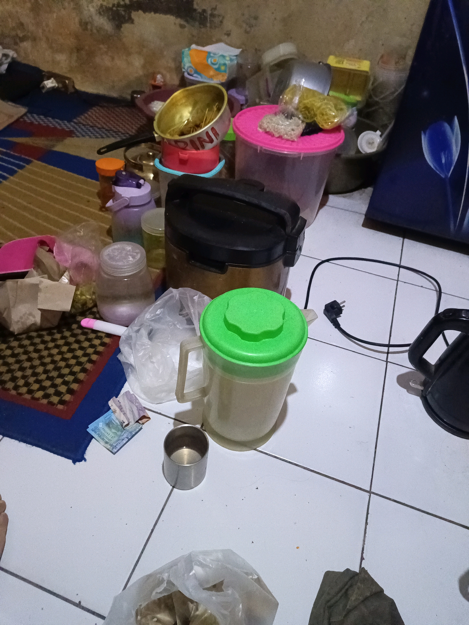 The image depicts a cluttered floor in what appears to be a residential interior in South Jakarta, Indonesia. The floor is tiled with white and grey squares, with visible grout lines. Various objects are scattered across the floor, suggesting a disorganized living space.
Prominently featured is a large, translucent pitcher with a bright green lid, filled with a light-colored liquid. Next to it, a black electric kettle with a handle is visible, its power cord plugged into an outlet on the wall. To the left of the pitcher, a small metal cup sits on the floor, containing a small amount of liquid. In front of the cup are several Indonesian Rupiah banknotes, folded and scattered.
Other items include several plastic containers of varying sizes and colors, including a pink-lidded container filled with yellow-colored objects, possibly food items. A gold-colored pot or bowl with red lettering is stacked on a pink container. There are also various bottles and jars, some containing liquids and others appearing empty. Several plastic bags, some containing objects, are also strewn about. A rolled-up rug with a checkered pattern is visible in the background, along with a dark-colored item that could be fabric. The walls in the background are rough and textured, with some dark discoloration. The overall impression is one of domestic disarray.