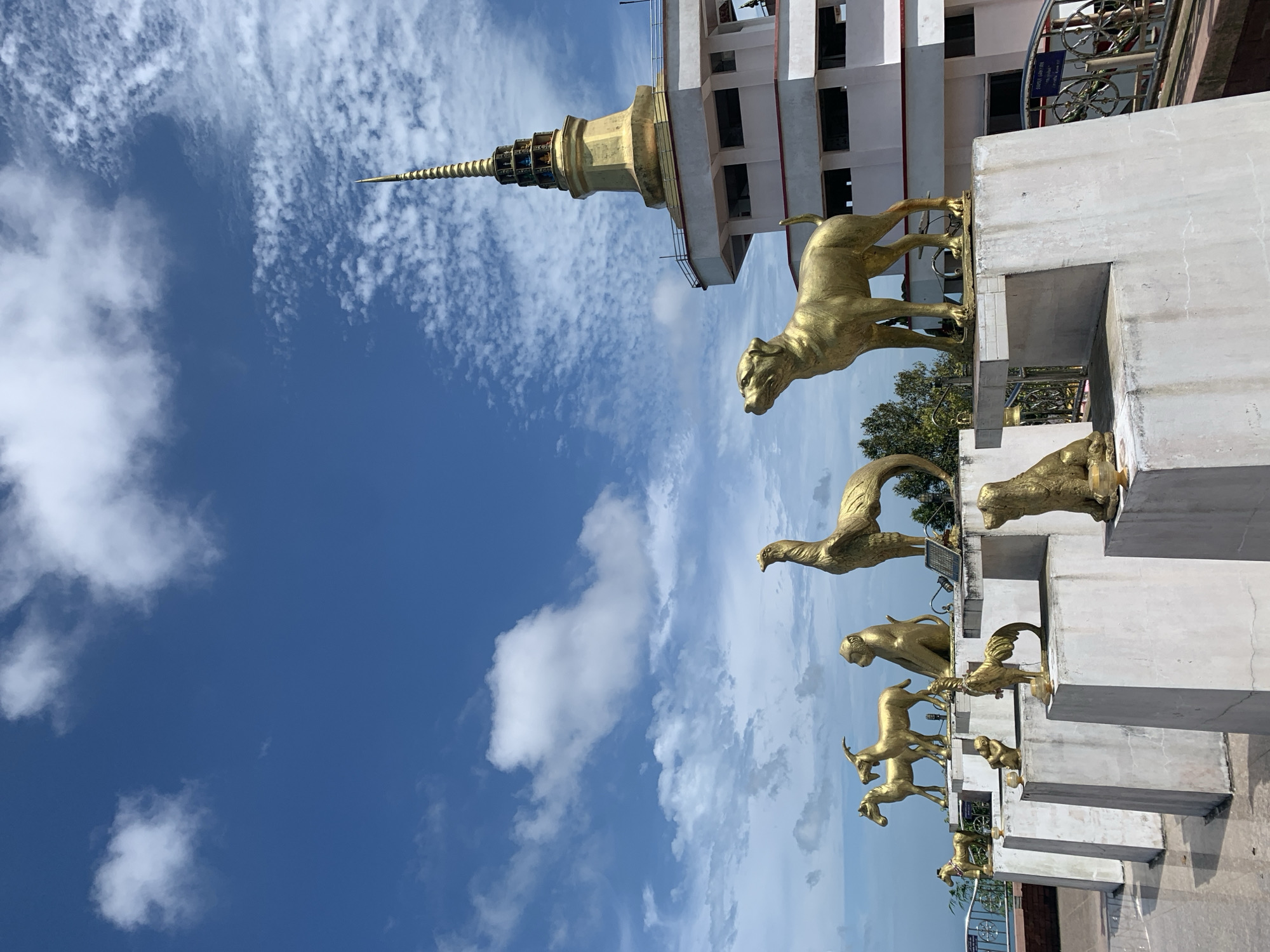 A group of golden animal statues stand in front of a tiered white and gold structure, possibly a temple, under a bright blue sky with scattered white clouds. The statues, depicted in various poses, appear to represent figures from the Chinese zodiac, including a dog, goat, monkey, and rooster. The architectural structure features a tall, golden, spire-like top, characteristic of some Buddhist temples. In the background, hints of greenery suggest a location with natural surroundings, possibly on a hill or elevated area, overlooking a body of water. The overall scene conveys a sense of cultural and religious significance, set against a scenic, sunny backdrop in Ko Samui District, Thailand.