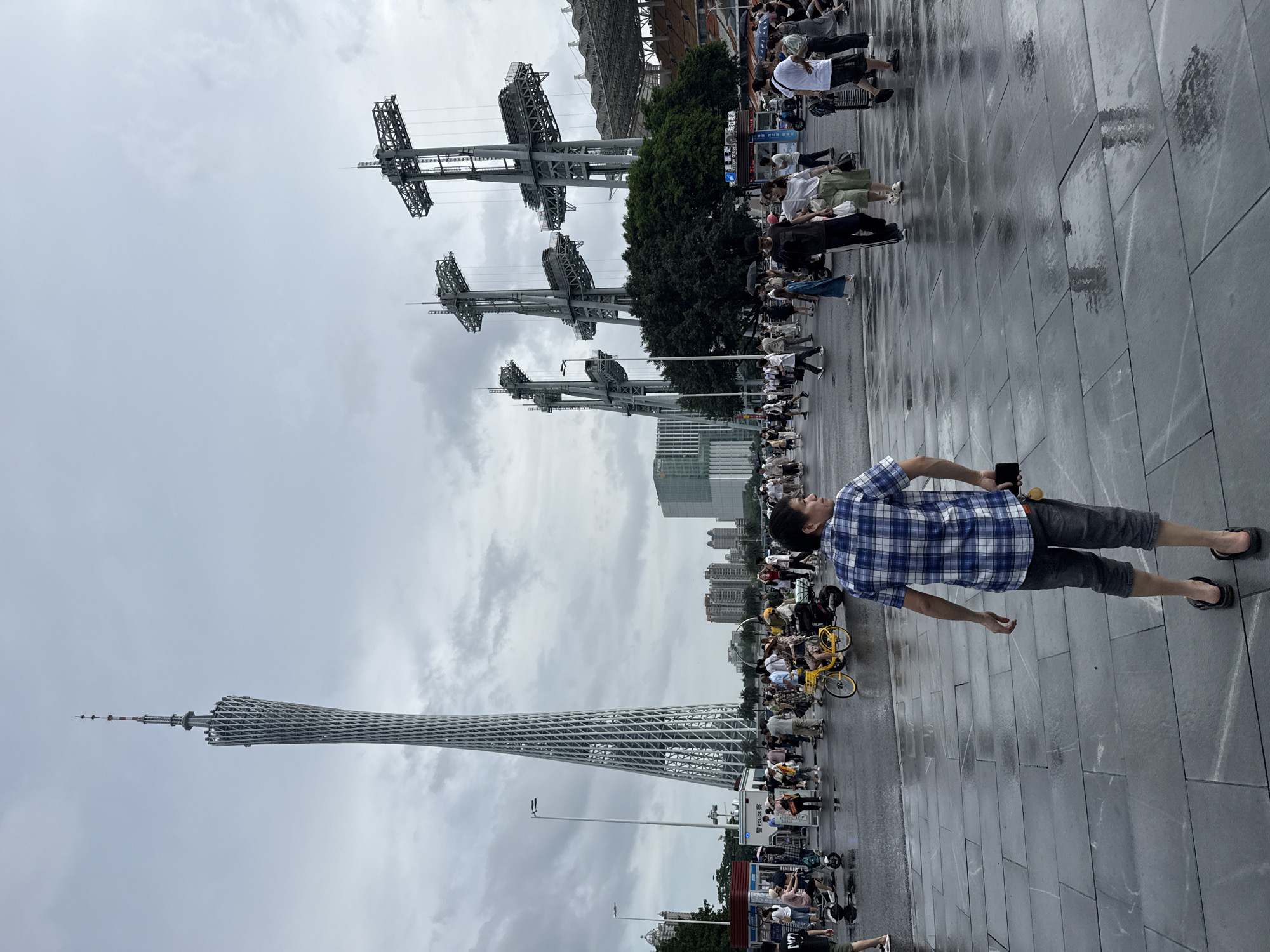 The image captures a bustling scene in the Tianhe District of Guangzhou, China, with the Canton Tower prominently featured in the background. The sky is overcast, and the ground, paved with large, dark tiles, is wet, suggesting recent rain. A diverse crowd of people is present, some walking, others on bicycles or electric scooters. Many are dressed in casual summer attire. The Canton Tower, a distinctive landmark, stands tall to the left, its intricate lattice structure visible. To the right, three large, industrial-looking structures with suspended platforms dominate the mid-ground, possibly part of a stadium or entertainment complex. In the foreground, a man wearing a blue and white checkered shirt and dark shorts is walking away from the camera, holding a dark mobile phone in his right hand. The overall atmosphere is one of lively urban activity under a subdued, cloudy sky.