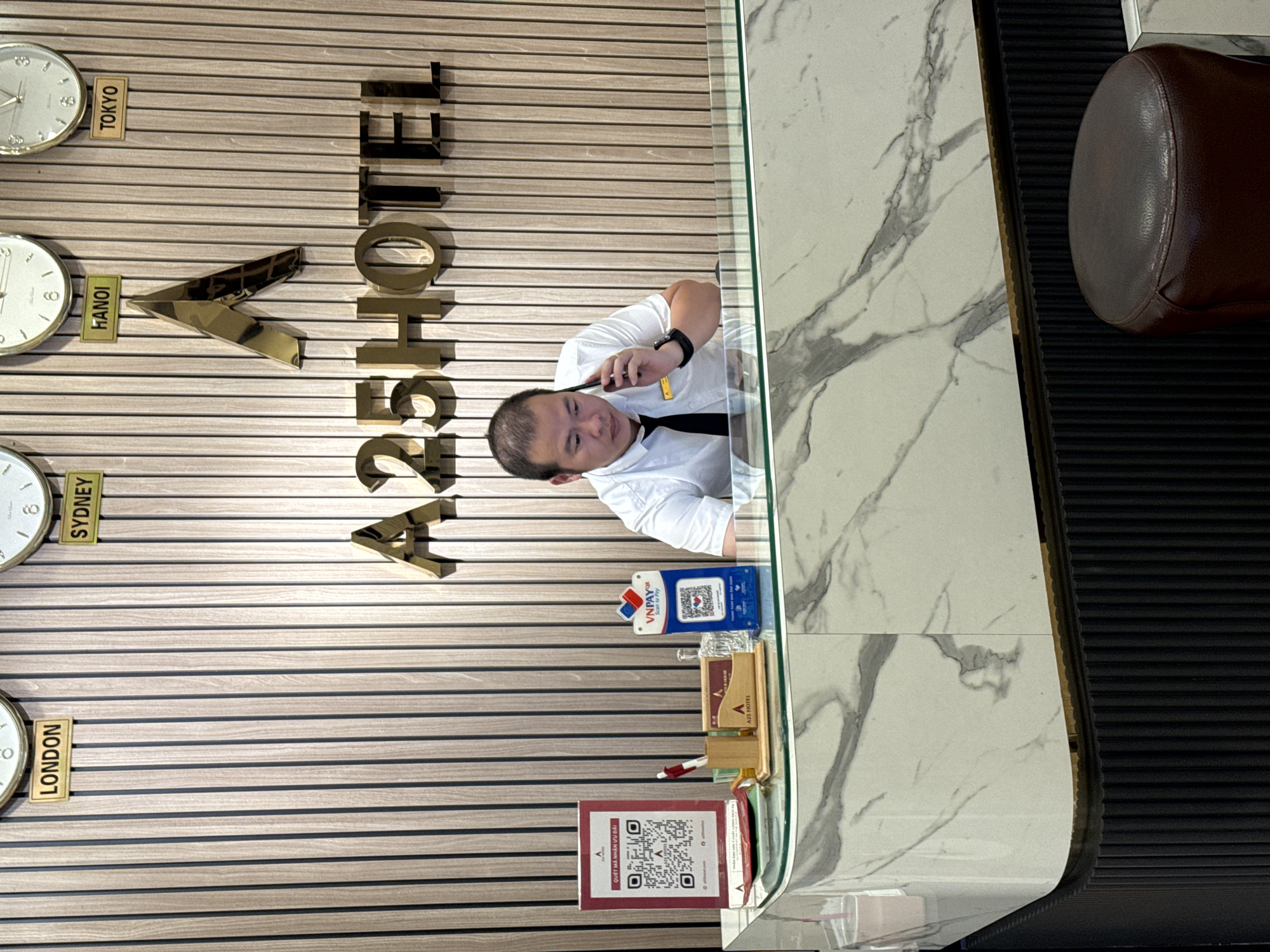 This media file captures a view of a hotel reception area in Hanoi, Vietnam. The focal point is a man, likely a hotel employee, wearing a white shirt and black tie, seen from the chest up. He is holding a phone to his ear, engaged in a conversation, with a slight tilt of his head.
Behind him, a wall is decorated with horizontal wooden slats, on which large, golden, three-dimensional letters spell out "A25 HOTEL". To the left of the hotel name, a series of clocks are mounted on the wall, each labeled with a city name: Tokyo, Hanoi, Sydney, and London, suggesting a global hospitality focus. These clocks are white and circular, with visible hands.
In front of the man is a glass-topped reception desk. On the desk, several items are visible: a blue VNPAY QR code display, a wooden pen holder with pens, and a red QR code display with Vietnamese text that translates to "Scan the code to receive discounts". There is also a small wooden holder with "A25 HOTEL" branding. The desk's surface has a marble-like pattern. To the right of the desk, a dark, ribbed material forms a partition or seating area, with a section of a brown leather seat visible. The overall impression is that of a modern, professional hotel reception environment. The lighting suggests it is daytime.