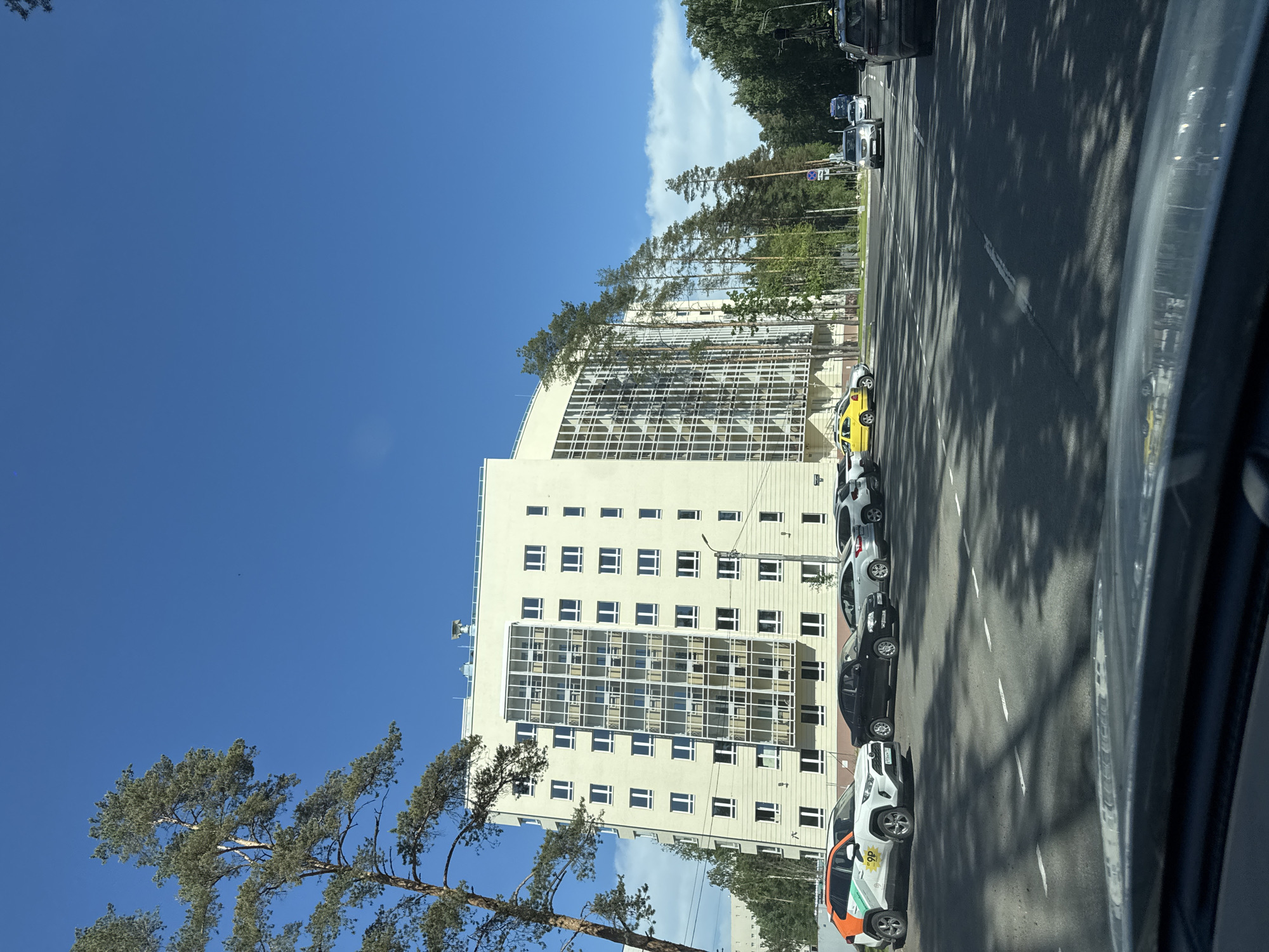 This image captures a street scene on a clear, sunny day in Pesochny, Russia. The perspective is from inside a vehicle, looking forward through the windshield.
In the left foreground, a tall pine tree with green needles and a brown trunk partially frames the scene. Behind it, a light-colored, multi-story building, likely a residential complex, dominates the left midground. This building features numerous rectangular windows with white frames and a prominent, multi-story glass-enclosed section on its facade. The rooftop has visible ventilation or communication equipment.
Adjacent to the building, a paved area serves as a parking lot, where several vehicles are parked. In the immediate left foreground, a white Toyota RAV4 SUV with orange and green "ОР" branding and partially visible Russian license plate (e.g., "У105ОМ 198") is parked. A black sedan, a silver sedan, and another white SUV are parked further along.
The central and right portions of the image are occupied by an asphalt road with visible white dashed lane markings. Strong, elongated shadows from unseen trees or structures are cast across the road and parking area. Traffic is present in the distance, with a silver SUV and a white sedan visible in the right lane, heading away from the camera. A blue truck is also visible further down the road. On the extreme right, a dark SUV is parked on the shoulder, facing away from the viewer.
A dense line of tall pine and deciduous trees borders the right side of the road, extending into the background. A single street light pole is visible between the building and the treeline. The sky above is a clear, deep blue with a few small, wispy white clouds visible in the upper right. No individuals are clearly discernible in the image.