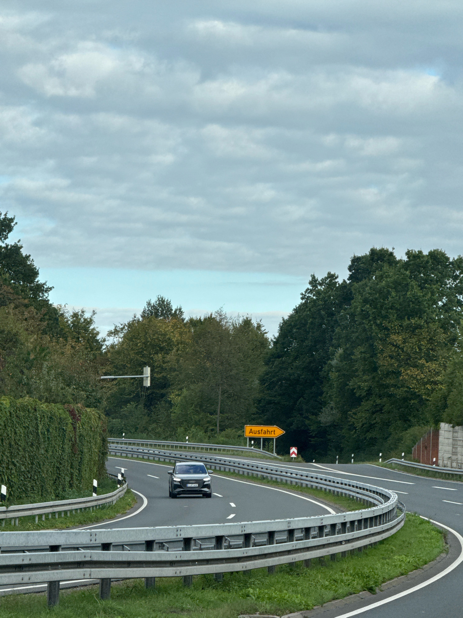 The image shows a two-lane road with a concrete barrier and metal guardrails on the right side, curving to the left. A dark colored car is traveling on the road. A yellow sign with the word "Ausfahrt" and an arrow pointing to the right is visible in the distance, indicating an exit. The road is bordered by lush green trees on both sides, and the sky is filled with overcast clouds. The scene appears to be on a highway or a major road outside of a city. While Herzberg, Germany, has a highway passing through it, the specific location cannot be determined from the image alone. There are no people visible in the image besides the occupant(s) of the car.