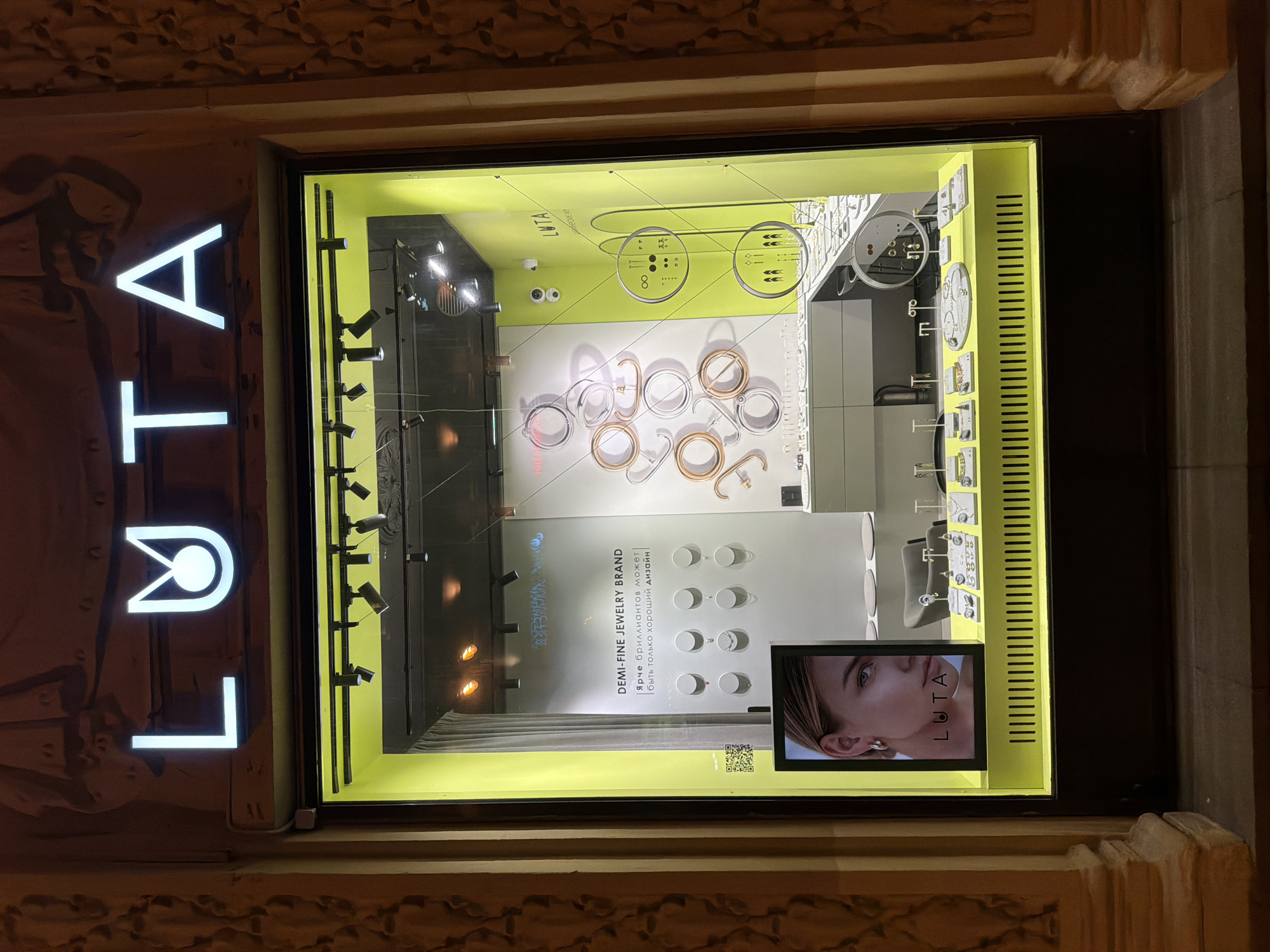 A nighttime view captures a brightly lit jewelry store window, revealing the brand "LUTA" in prominent vertical signage. The window display features an array of jewelry, including rings and earrings, neatly arranged on white and green display boards. A digital screen in the window shows a close-up portrait of a woman wearing jewelry, with the "LUTA" logo visible. Text on a display board describes the brand as "DEMI-FINE JEWELRY BRAND" and includes a Russian phrase that translates to "Brighter than diamonds can only be good design." The store's exterior is framed by ornate stone architecture, typical of urban buildings, suggesting a location within Moscow, Russia. Security cameras are visible on the wall of the window display, suggesting surveillance.