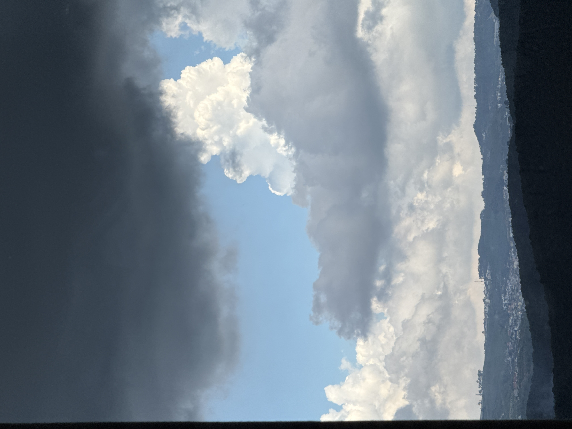 The image shows a cloudy sky over a hilly landscape with a town nestled in the valleys. The sky is filled with various types of clouds, ranging from dark, heavy storm clouds to bright, puffy white cumulus clouds. In the distance, rolling hills covered in dark green foliage form the horizon. A town with buildings of various sizes and colors is visible on the slopes of the hills, with some structures appearing to be houses and others possibly commercial buildings. The overall impression is of a serene, natural environment with a human settlement integrated into the landscape. The specific location in Đa Lộc, Vietnam, is not explicitly identifiable from the image alone, but the terrain and vegetation suggest a tropical or subtropical climate.
