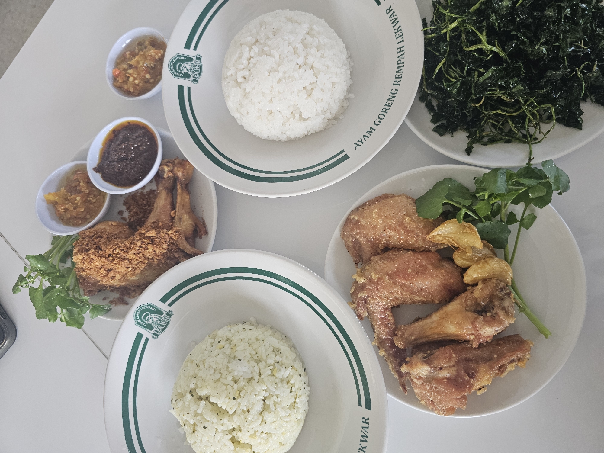 A top-down, slightly angled shot captures a meal spread out on a white, tiled surface. The meal consists of several dishes, primarily featuring fried chicken and rice, with accompanying condiments and vegetables.
In the upper center, a plate holds a generous portion of fried chicken, coated in crispy crumbs and appearing seasoned. Beside it, a small plate is laden with three small bowls of chili paste or sambal, varying in color from light orange to a deep reddish-brown. A sprig of fresh green leaves, possibly watercress, garnishes this plate.
To the left, a white plate with green trim showcases a mound of fluffy white rice, speckled with what appear to be herbs. The same branded plate with green trim is visible on the right, also holding a portion of white rice.
Below these, another white plate displays an assortment of fried chicken pieces, including wings and drumettes, alongside some fried garlic. A pile of dark green, wilted leafy vegetables is presented in a separate white dish on the lower right.
The branding on the plates, "AYAM GORENG REMPAH LEKWAR," suggests this is a dish from a restaurant or food stall named Lekwar, specializing in spiced fried chicken. The overall impression is of a hearty and flavorful Indonesian meal, ready to be consumed. The location is confirmed to be Banyuurip, Indonesia, based on the context provided.