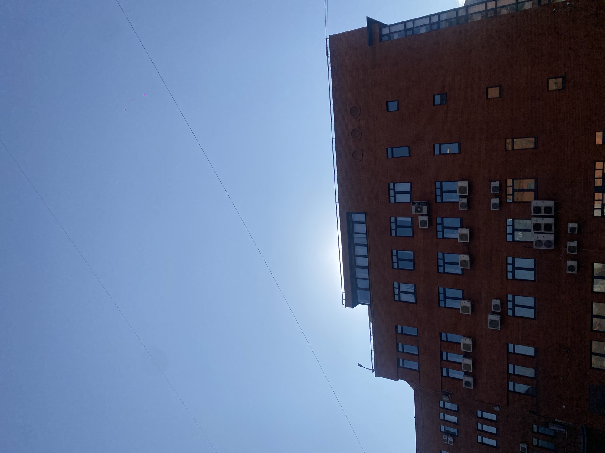The image displays a multi-story building, likely commercial or mixed-use, with a reddish-brown facade, under a clear, light blue sky. The sun is positioned directly behind the building's upper section, creating a bright halo effect and partially silhouetting the structure. Numerous rectangular windows with dark frames are visible across the building's face, arranged irregularly. White outdoor air conditioning units are mounted beneath many of the windows on various floors.
The roofline includes a section with a dark railing and a light fixture resembling a streetlamp on the far left. A setback upper floor features a band of horizontal windows. To the right, three distinct circular indentations are visible on the upper facade. The building's right side incorporates a multi-story glass facade with a dark grid-like framework. Two thin, dark wires extend diagonally across the upper portion of the sky. Three faint, magenta-colored specks are present in the upper right sky area, consistent with photographic sensor dust or lens artifacts. No people or specific identifying landmarks are discernible to pinpoint the exact location within the city of Moscow, Russia.