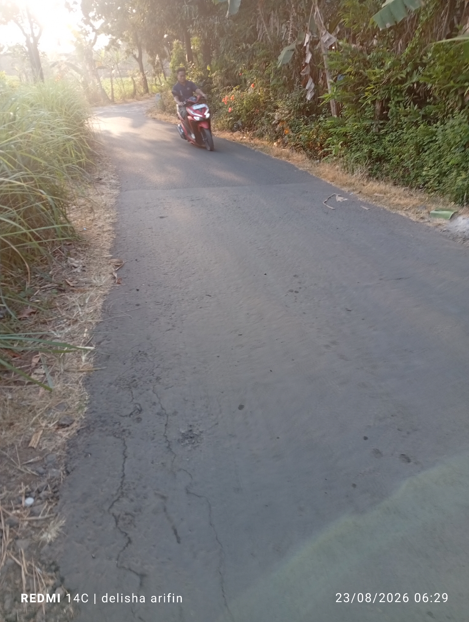 The image shows a road in Jati Gunting, Indonesia, with a person riding a red motorcycle on the left side of the road. The road is paved and appears to be in good condition. There are trees and vegetation on both sides of the road. The sky is bright and sunny, suggesting it is daytime. The timestamp in the bottom right corner indicates the image was taken on August 23, 2026, at 06:29. The text "REDMI 14C | delisha arifin" is visible in the bottom left corner, likely indicating the phone model and the photographer's name.
