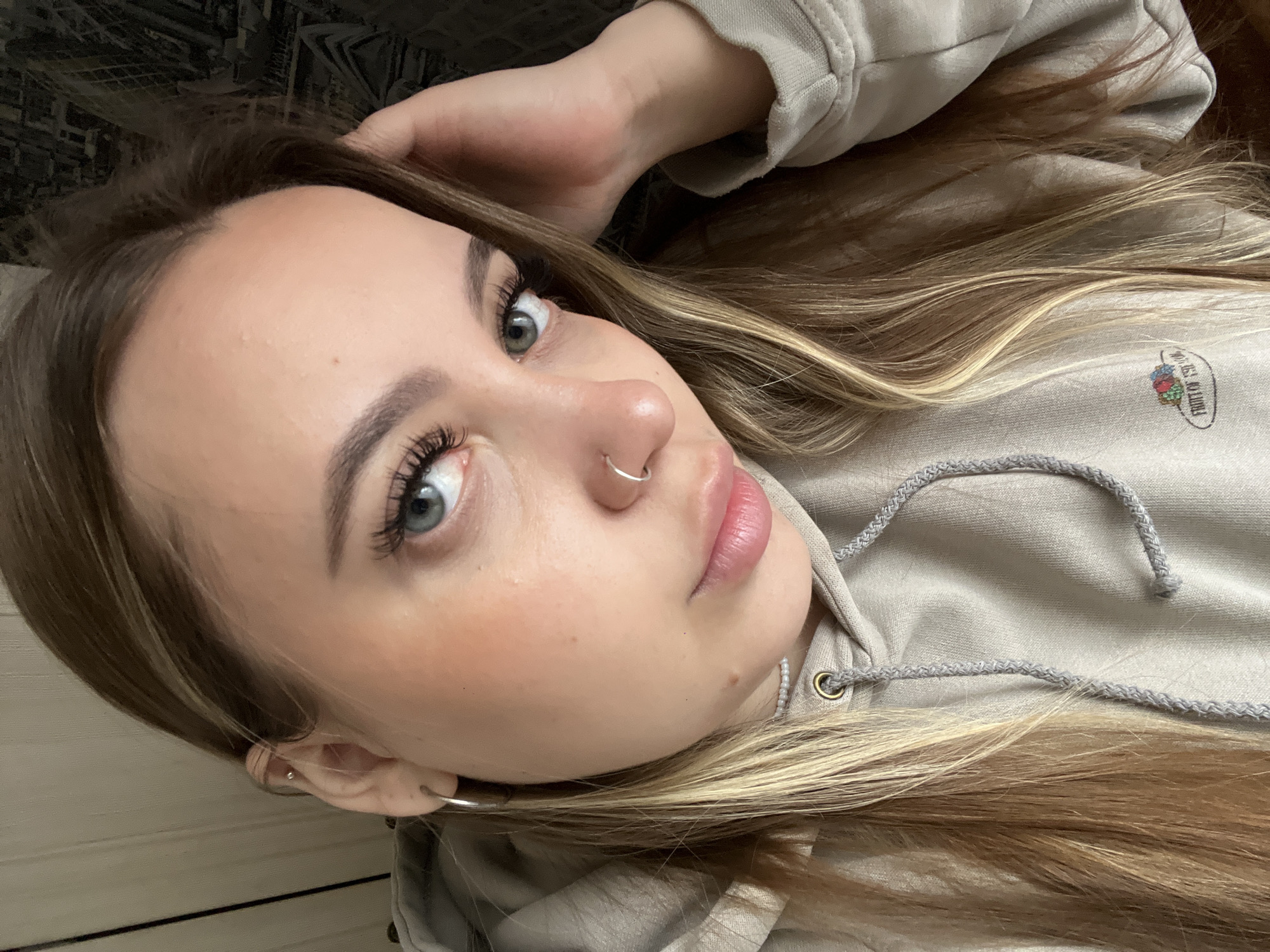 The image is a close-up, angled portrait of a young woman looking directly into the camera. Her head is tilted, giving a casual and relaxed pose. She has long, light brown hair with blonde highlights, which flows around her. Her eyes are a striking blue-grey, accentuated by long, dark eyelashes. Her eyebrows are well-defined, and her skin appears fair with a subtle flush on her cheeks.
The woman wears a silver hoop nose piercing and two ear piercings: a silver hoop in her earlobe and a small stud in her helix. Her lips are naturally pink. She is dressed in a light beige or light grey hooded sweatshirt, with visible grey drawstrings around the neck. On the upper right side of the hoodie, near her shoulder, a colorful "FRUIT OF THE LOOM" logo is clearly visible. A delicate, pearl-like necklace can also be seen around her neck.
The background on the left is dark and indistinct, possibly a patterned fabric, while the lower right shows a lighter, textured surface, suggesting an indoor setting. The lighting appears soft, indicating an interior environment. The overall mood of the image is calm and relaxed, capturing a personal moment.