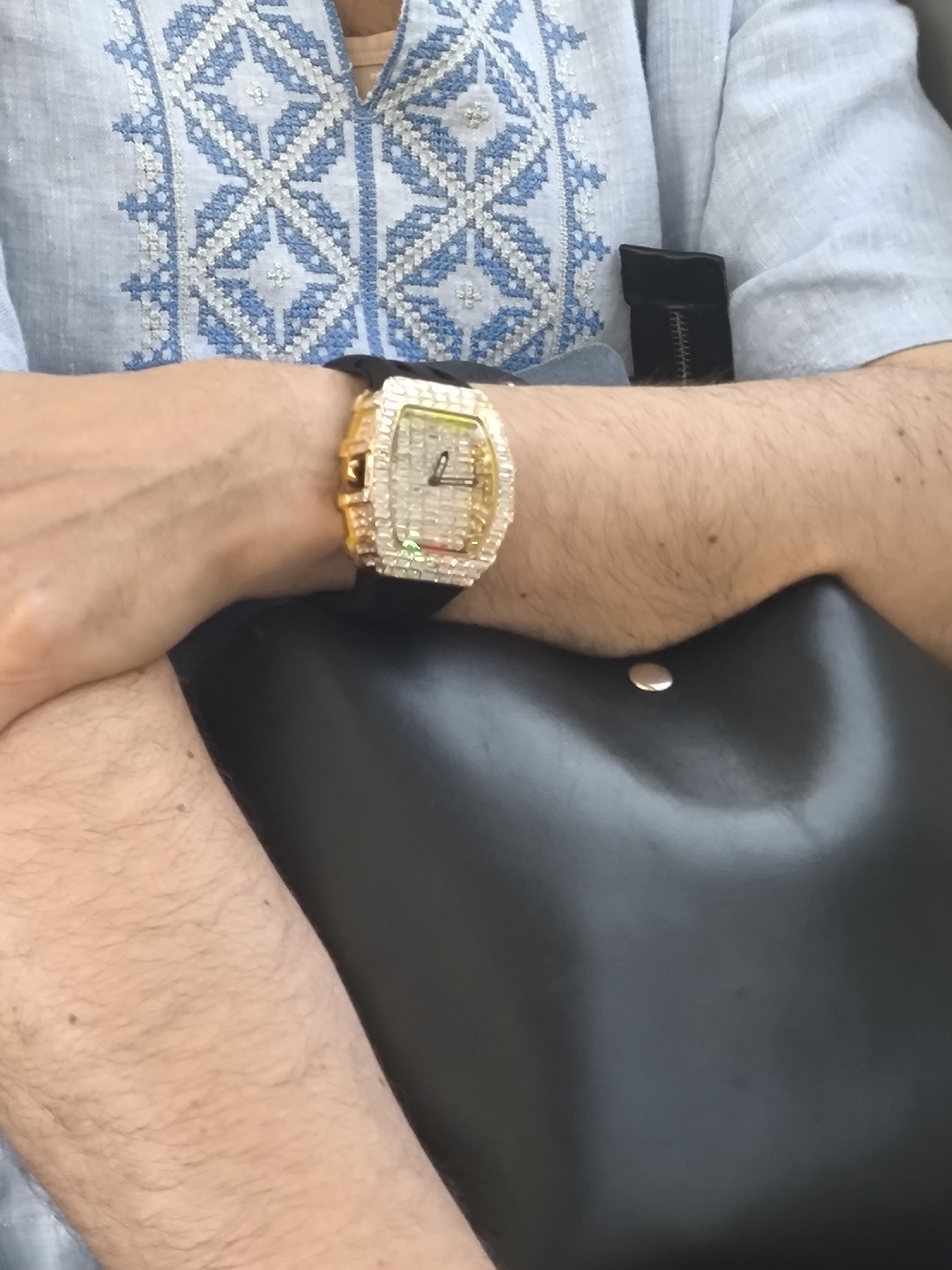 A person's arm rests on a black leather surface, with a gold-toned, diamond-studded watch visible on their wrist. The person is wearing a light blue shirt with a white and blue embroidered pattern. A black object is visible behind the arm and shirt. The location is unknown.