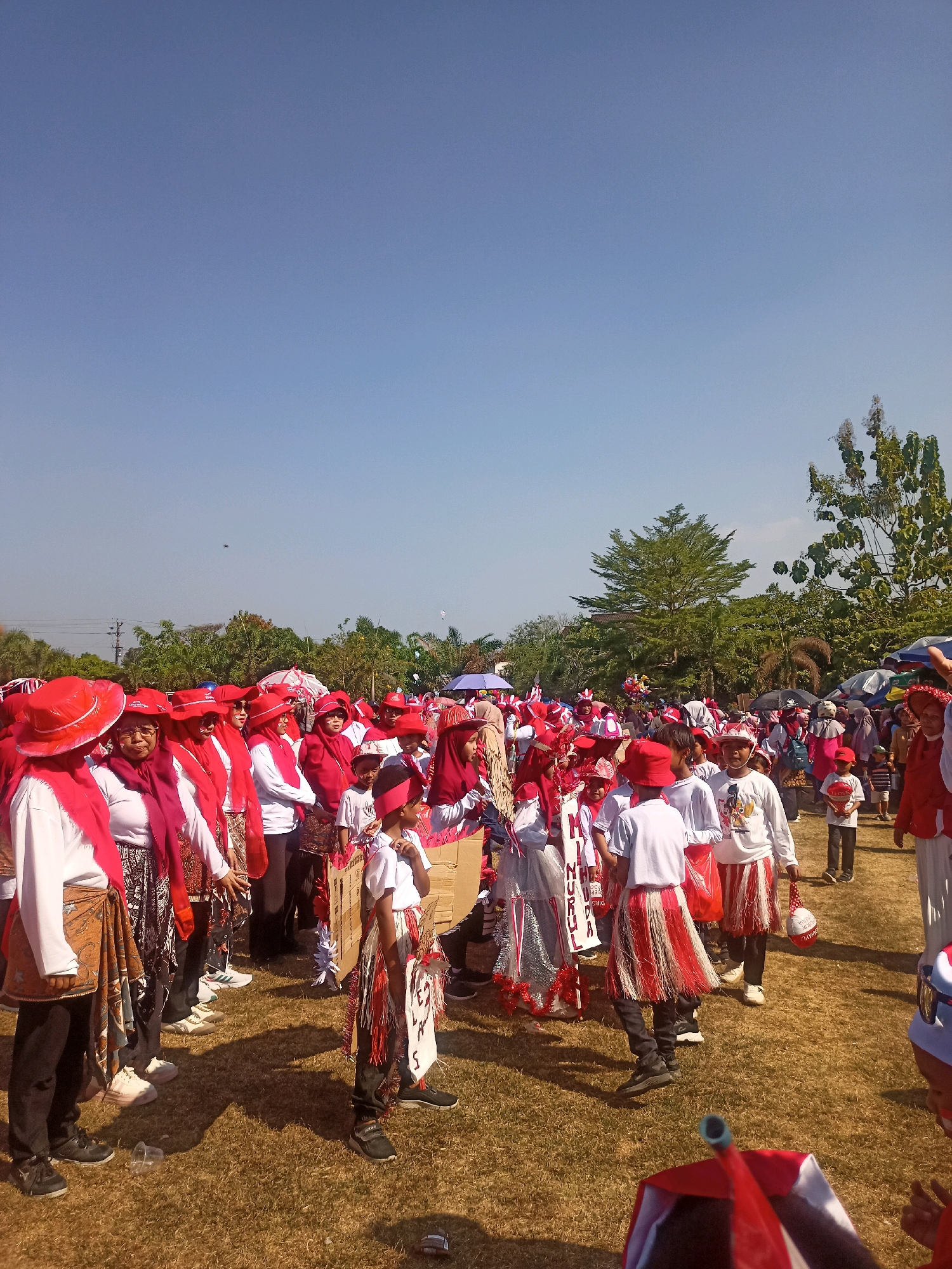 The image depicts a daytime outdoor scene in Sumberlawang, Indonesia, featuring a large group of people, predominantly children and adults, dressed in predominantly red and white attire. The gathering appears to be a parade or cultural event. Many participants are wearing red hats, red scarves, and clothing with red and white patterns. Some individuals are adorned with traditional or festive costumes, including striped skirts and elaborately decorated headwear. The ground is covered in dry grass, suggesting an open field or park. In the background, trees and some structures are visible, along with umbrellas and tents. The sky is clear and blue, indicating sunny weather. The people are congregated in a loose formation, with some walking and others standing. The overall atmosphere suggests a community event or celebration.