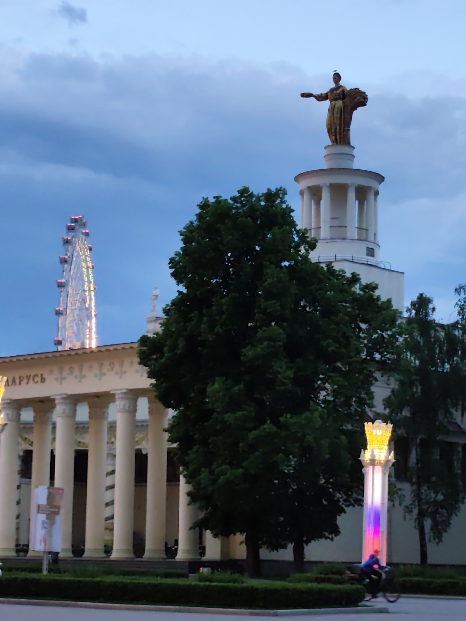The photograph depicts a street scene at dusk in Moscow, Russia, featuring several prominent landmarks. A brightly lit Ferris wheel is visible in the background. A large building with white pillars and the word "БЕЛАРУСЬ" is in the middle ground. A statue of a woman holding what appears to be a bundle of wheat stands atop a white tower. A large tree obscures part of the view. In the foreground, a person on a bicycle rides past a decorative illuminated pillar.