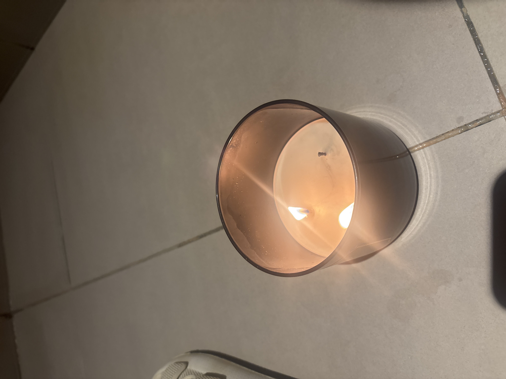 The image shows a lit candle in a glass container, placed on a tiled floor. The candle has two visible flames and a partially melted wax surface. To the left of the candle, a portion of a white sneaker is visible. The background consists of light-colored tiles and grout lines. There is no indication of the location being in Dubai, United Arab Emirates, or any other specific city. The scene is static and does not depict any active interactions or events.