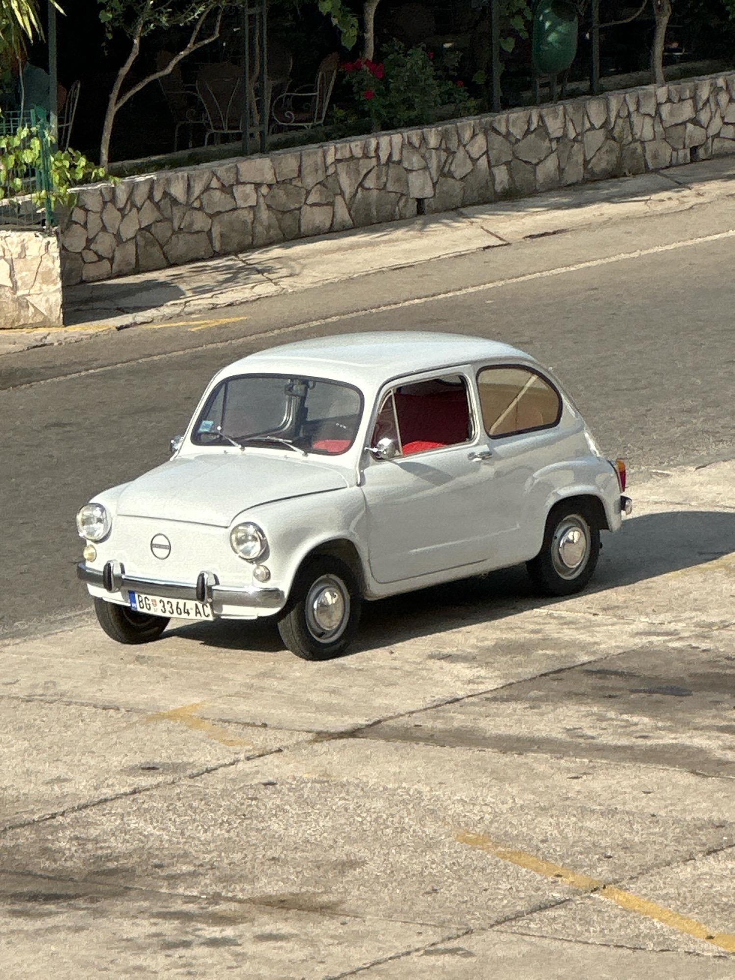 A vintage white Fiat 600 is parked on a concrete surface next to a street. The car has a red interior and a license plate that reads "BG 3364 AC". In the background, a stone wall separates the street from an area with greenery and outdoor seating. The scene appears to be in Budva, Montenegro, given the license plate format. The lighting suggests it is daytime.