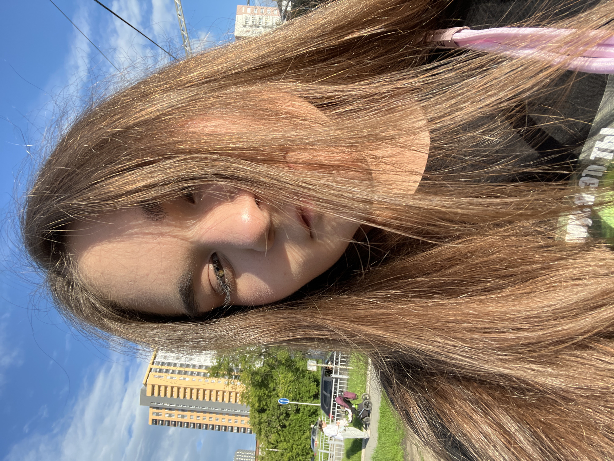The image is a selfie taken outdoors in the city of Yekaterinburg, Russia. The foreground is dominated by the face and long, light brown hair of a young person, partially obscuring their features. The background shows a typical urban environment with tall residential buildings, green trees, a sidewalk, a road with parked cars, and utility poles against a blue sky with scattered clouds. To the left, a woman is seen pushing a stroller, indicating a residential area or park. The lighting suggests it is daytime, with sunlight creating highlights and shadows on the subject's face and hair.