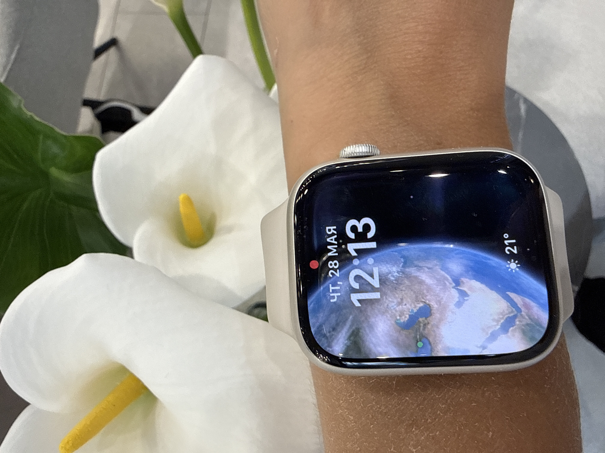A close-up shot shows a person's wrist wearing an Apple Watch with a world map face displaying the time as 12:13. The watch face also indicates that it is Thursday, May 28th, and the temperature is 21 degrees Celsius. The surrounding environment includes white calla lilies, suggesting a bright and possibly indoor setting. The location is inferred to be Sochi, Russia, based on the watch's use of Russian date and time formats.