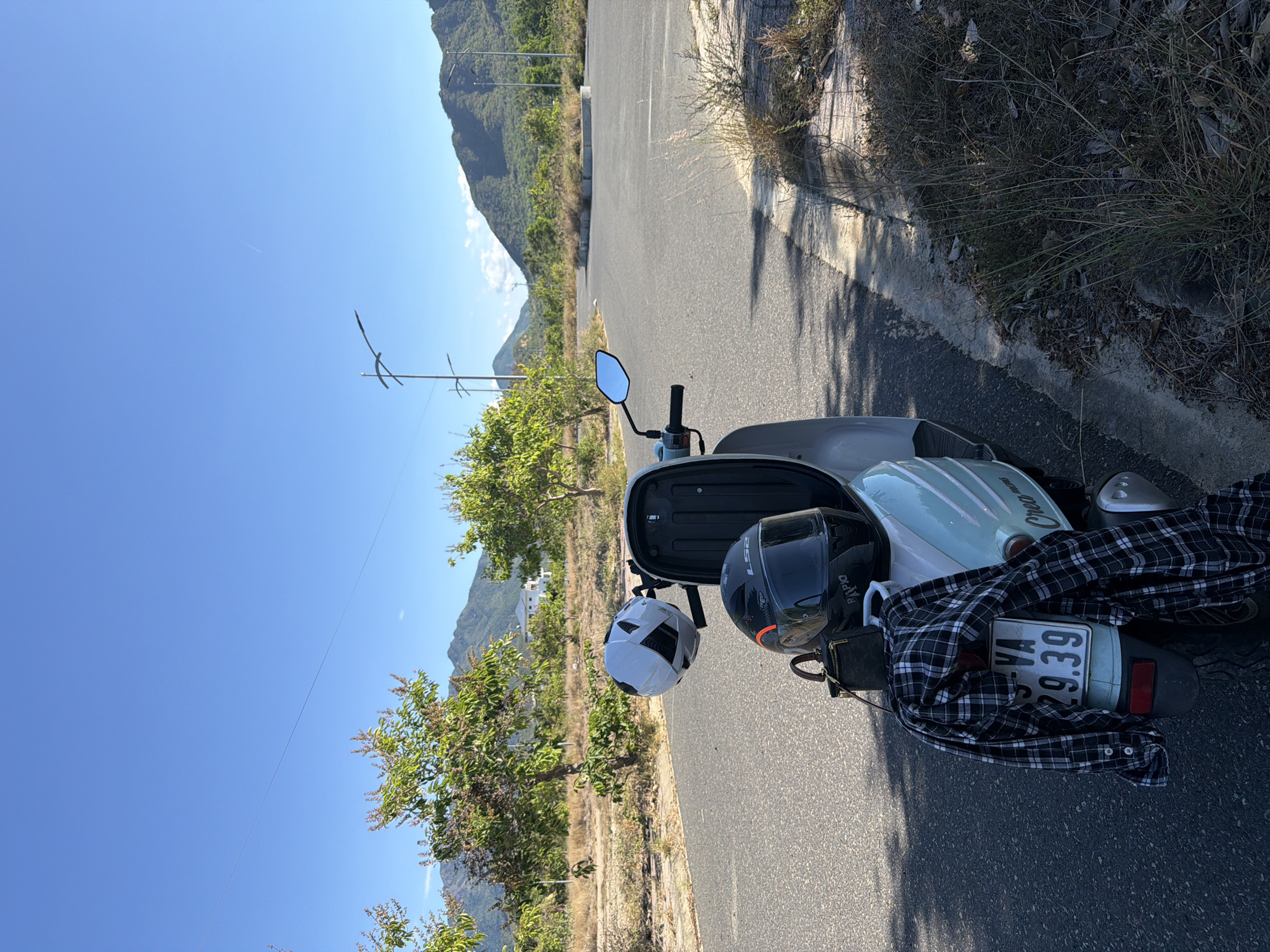 Image shows a scooter parked on a paved road with a view of mountains in Nam Nha Trang Ward, Vietnam. The scooter has two helmets, one black and one white, and a black and white plaid shirt draped over the back. The scooter's license plate is visible. The road has sparse vegetation on the sides, and overhead power lines are visible. The sky is blue.