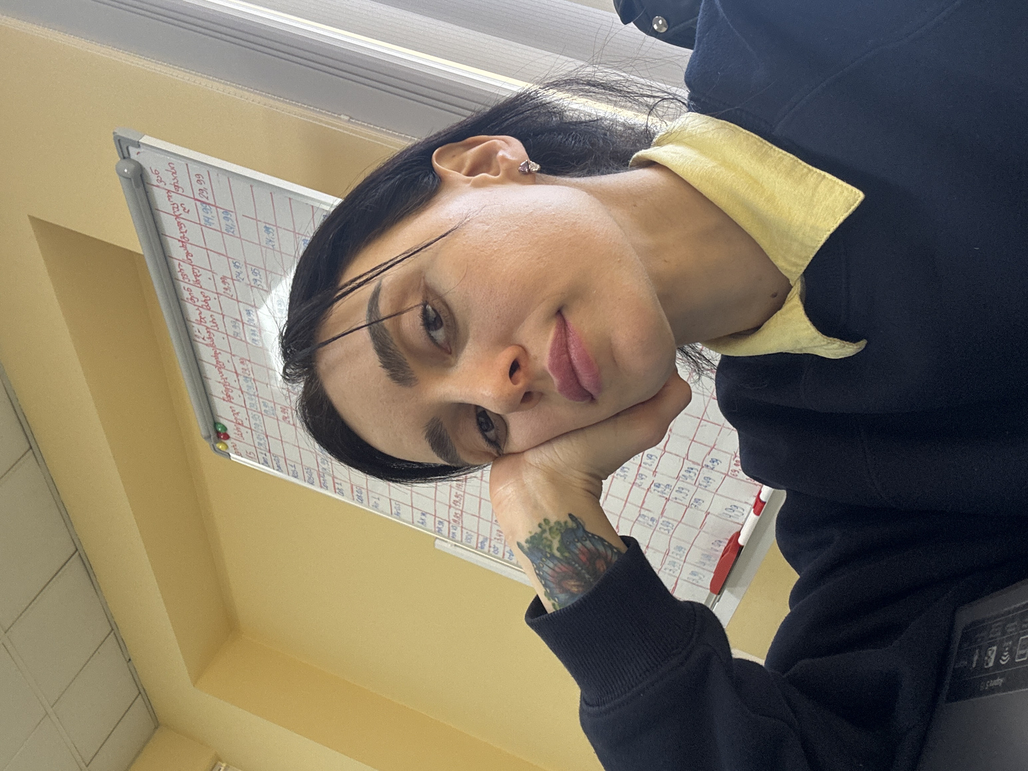A young adult female rests her face on her left hand, in front of a white board with red and black writing. She is wearing a black long-sleeved shirt with a yellow collar and a tattoo on her left wrist. The location is likely an office setting in Tbilisi, Georgia, based on the appearance of the whiteboard with text in Georgian. The lighting is bright and likely fluorescent.