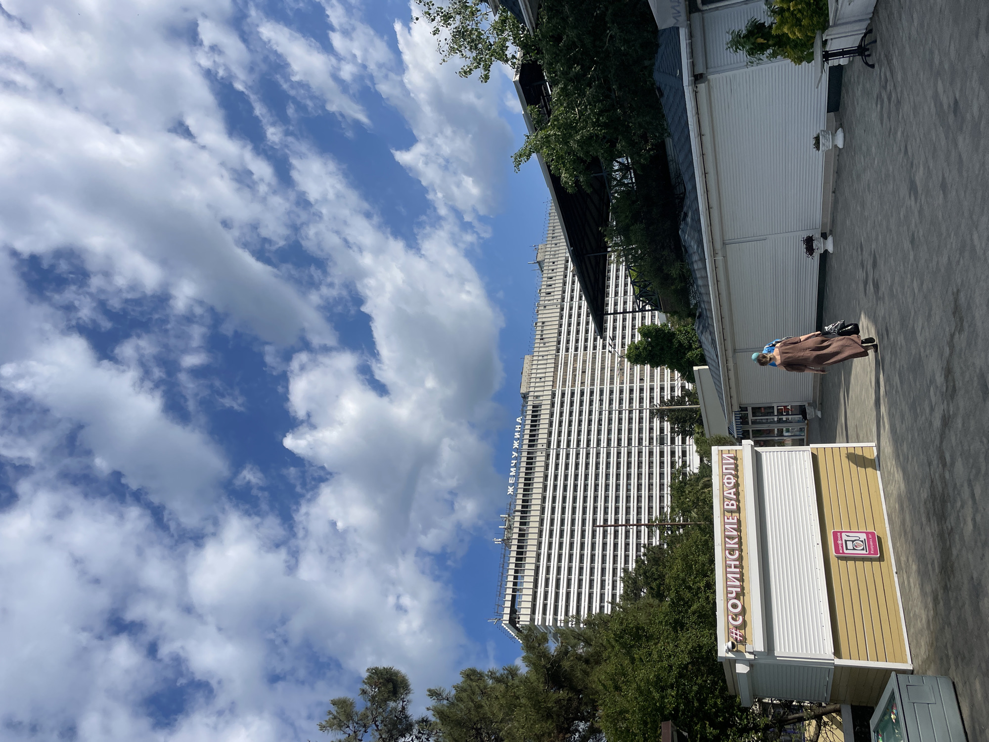 A medium, eye-level, full outdoor shot shows a scene near the Black Sea coast in Sochi, Russia. The image is rotated 90 degrees clockwise, with the sky at the left of the frame. A partly cloudy sky, with a rich blue color and numerous white clouds, is prominently displayed.
A tall, white, linear building stands against the blue sky. It is the "Zhemchuzhina" hotel. The hotel's name is written in blue, Cyrillic letters near the top of the building. The building's architecture is characterized by multiple stacked horizontal lines.
In the foreground, there is a light-brown kiosk with the word "#SOCHINSKIE VAFLI" written in pink on the front. A woman wearing a brown dress, blue headscarf, and black shoes stands near the kiosk. The woman is seen from the back. The location is a gray paved promenade.