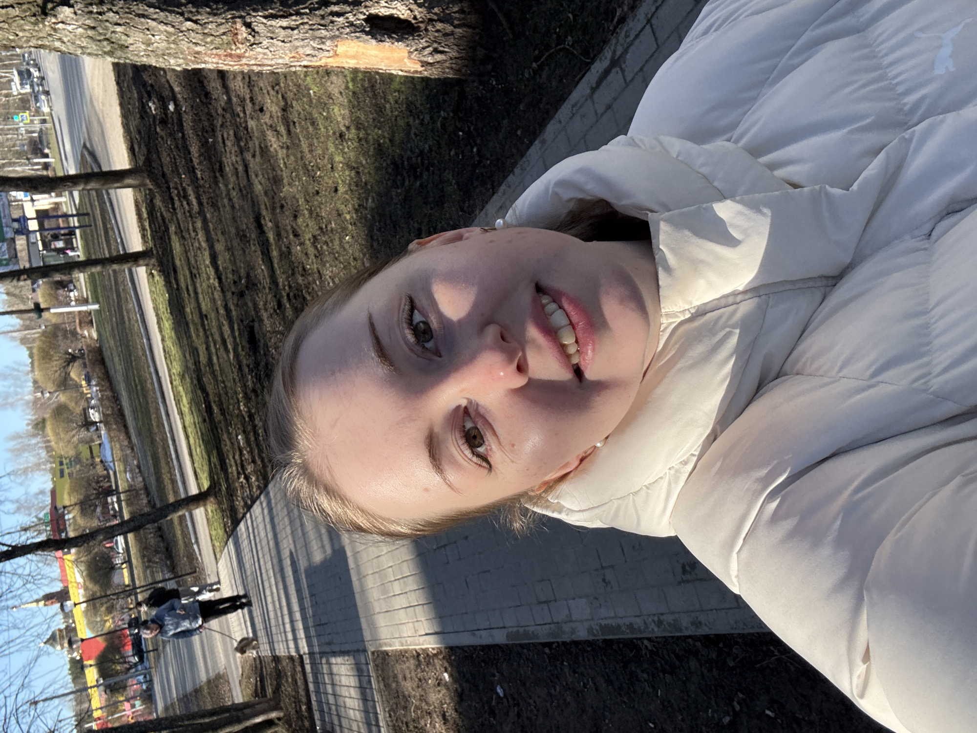 A young Caucasian woman with blonde hair and hazel eyes, wearing a white puffer jacket, smiles at the camera while standing on a paved path. The path is made of gray bricks and is partially shaded. To the left of the path is an area of dirt and grass with several bare trees. In the background, across a street, there are colorful buildings and a person walking a dog. A tree trunk is visible in the upper left corner of the frame. The photograph is taken at an angle, giving a slightly tilted perspective. The location is unknown.