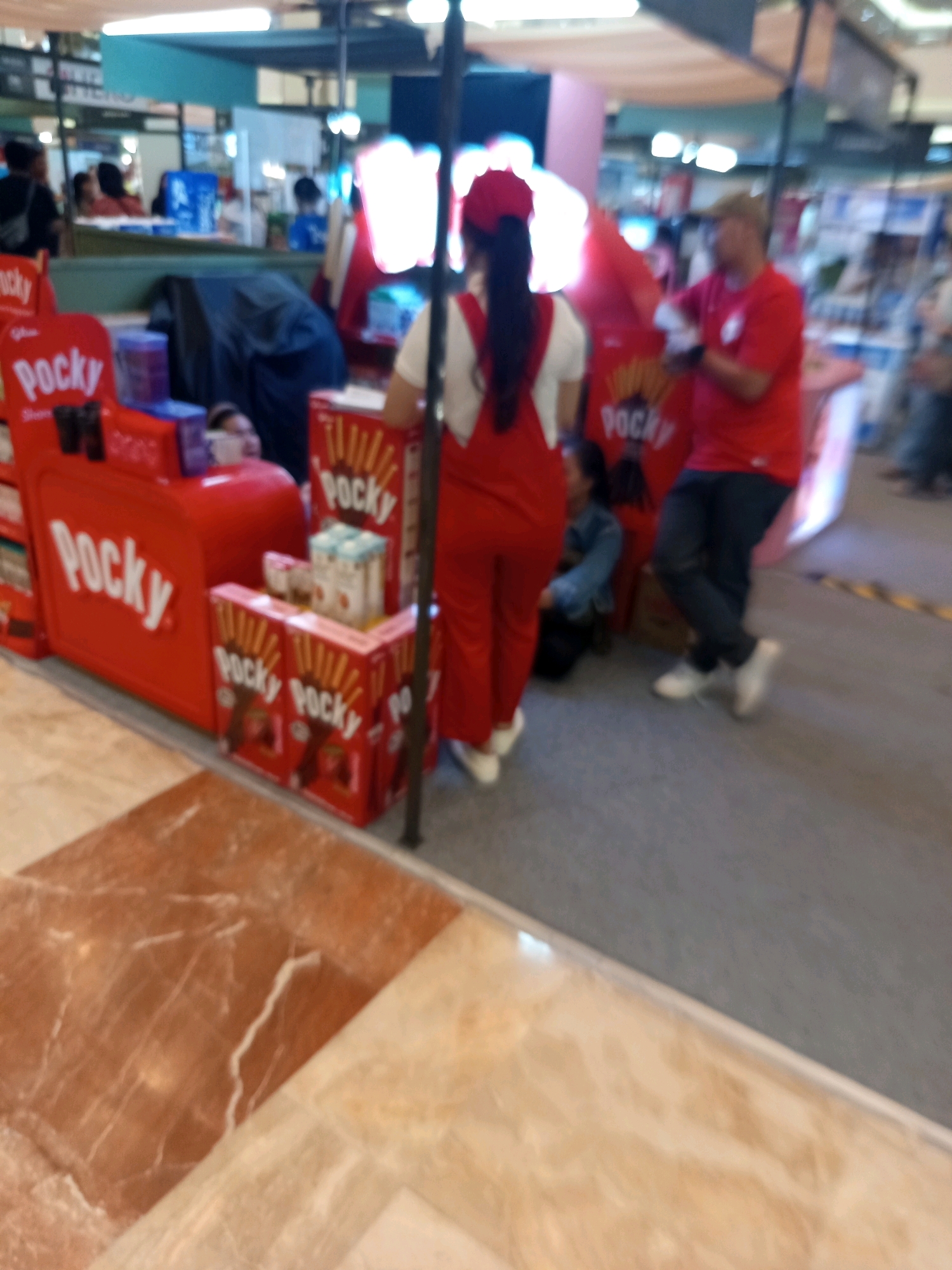 The image depicts a promotional stall for Pocky snacks, likely located in West Jakarta, Indonesia, given the context of Pocky's popularity there. Two individuals are prominently featured: a woman in red overalls and a red headband, and a man in a red t-shirt and a tan cap. The woman is standing and facing away from the camera, with her hands resting on a red display. The man is also facing away, leaning against a stall or display. Several Pocky boxes and containers are visible on the stall, in various sizes and designs. In the background, other individuals are present, some interacting with the stall and others moving about in what appears to be a busy commercial area or shopping mall. The floor has a tiled pattern of brown and beige marble. The lighting is bright and artificial, indicative of an indoor commercial setting.