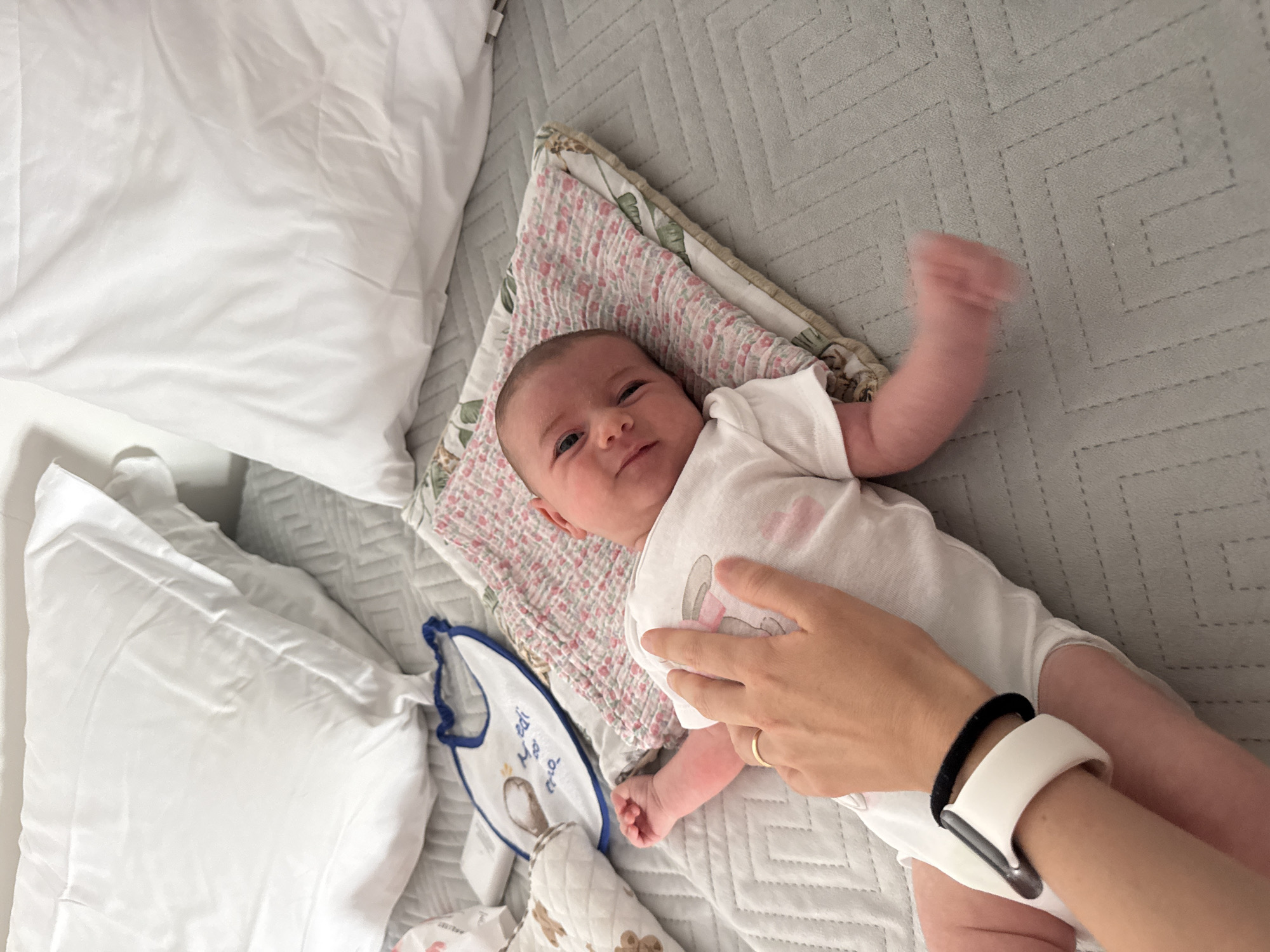 A newborn baby is lying on a bed with white pillows in the background. The baby is wearing a white onesie with a cartoon design and is lying on a small pink and white patterned blanket. A hand with a gold ring and a white smart watch is gently touching the baby's chest. A bib with a drawing of a rabbit and the text "Miudi" is on the bed to the left of the baby. The bed is covered with a grey textured blanket. The overall scene suggests a domestic setting, likely a bedroom, in Isola delle Femmine, Italy.