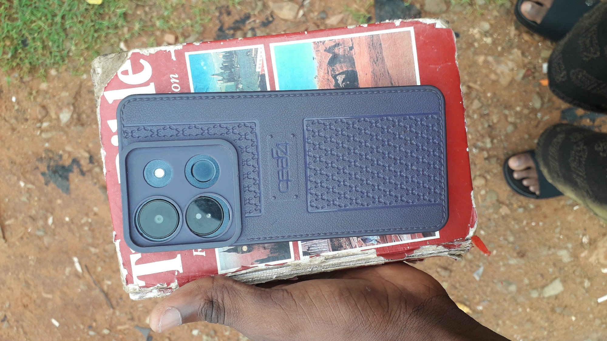 The image shows a hand holding a worn, red book with photographs affixed to its cover. A blue smartphone with a textured case, bearing the brand name "Zeec," rests on top of the book. The phone features a prominent camera module. Adjacent to the book and hand, a pair of feet in sandals are partially visible. The background consists of earth and sparse vegetation. The setting is Nkpor, Nigeria.