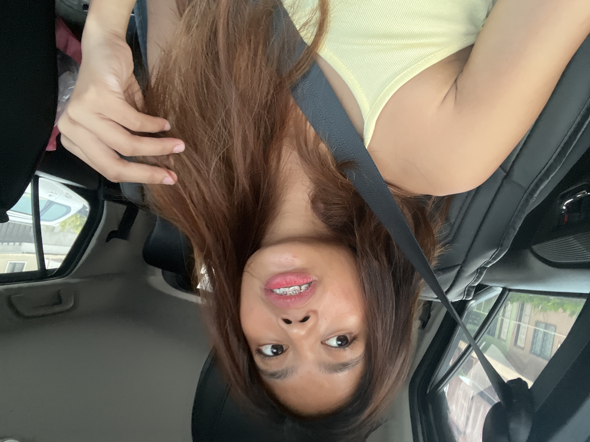 A young woman with brown hair and braces is sitting in the driver's seat of a car, looking to her left with her mouth slightly open. She is wearing a yellow tank top. The interior of the car is visible, including the black leather seats and a black seatbelt. Through the window on the left, houses with green roofs and white siding are visible, suggesting a residential area, potentially in Davao City, Philippines. A silver car is parked outside the window on the right. The woman's right hand is touching her hair.