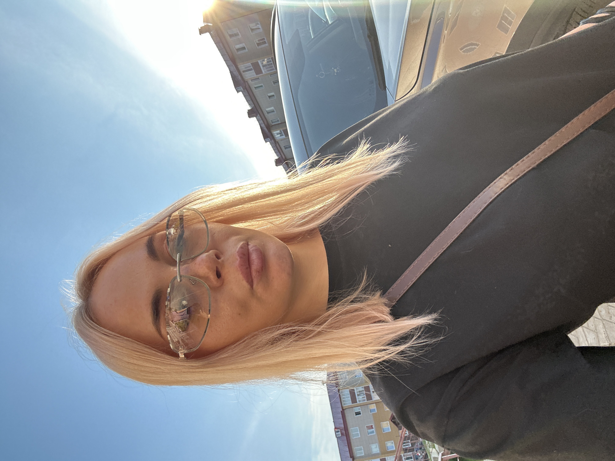 A blonde woman wearing sunglasses and a black t-shirt is taking a selfie outdoors. She is positioned in the foreground, with a car and a building in the background. The sun is visible in the upper right corner, creating lens flare. The sky is clear and blue. The woman's right shoulder is visible, and a brown strap from a bag crosses her chest. The building in the background appears to be an apartment complex. The scene is likely in a residential area of a city.