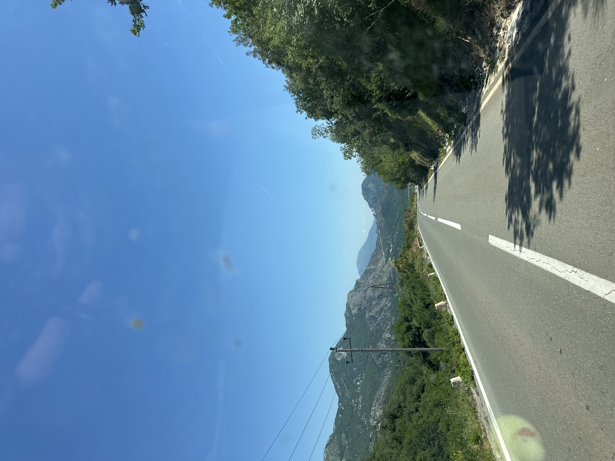 The image depicts a winding road in Novoselje, Montenegro, flanked by lush greenery and rocky mountainous terrain. The road is asphalt, marked with a white center line and an edge line. Utility poles with power lines run alongside the road on the left side. The sky is clear and blue. Shadows from trees are cast onto the right side of the road. The vegetation consists of trees and bushes growing along the roadside and on the slopes of the mountains.