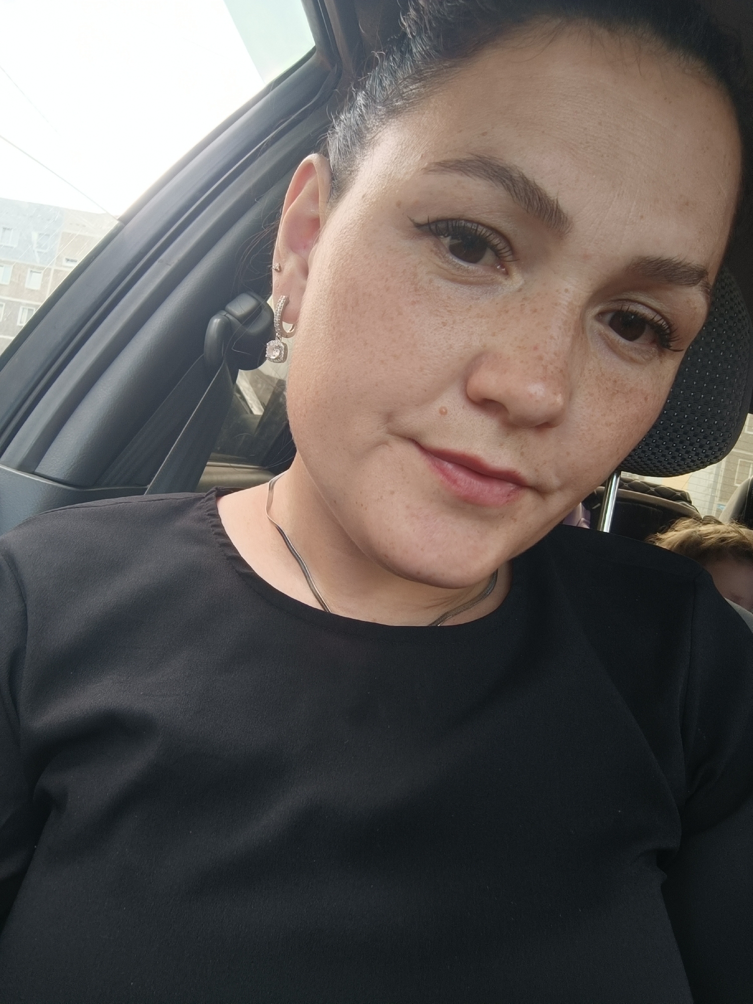 A woman with dark hair pulled back and numerous freckles on her face is positioned in the foreground of the image, looking slightly to the right. She is wearing a black shirt and a thin silver necklace. A square-shaped earring with a clear stone dangles from her left earlobe. The background suggests she is inside a vehicle, with a car seat visible behind her. Outside the vehicle, buildings and a bright sky are apparent, consistent with an urban environment. The overall lighting indicates it is daytime, likely outdoors. The city of Chaikovsky, Russia, is a residential area with numerous apartment buildings, suggesting this scene could have been captured in such a locale.