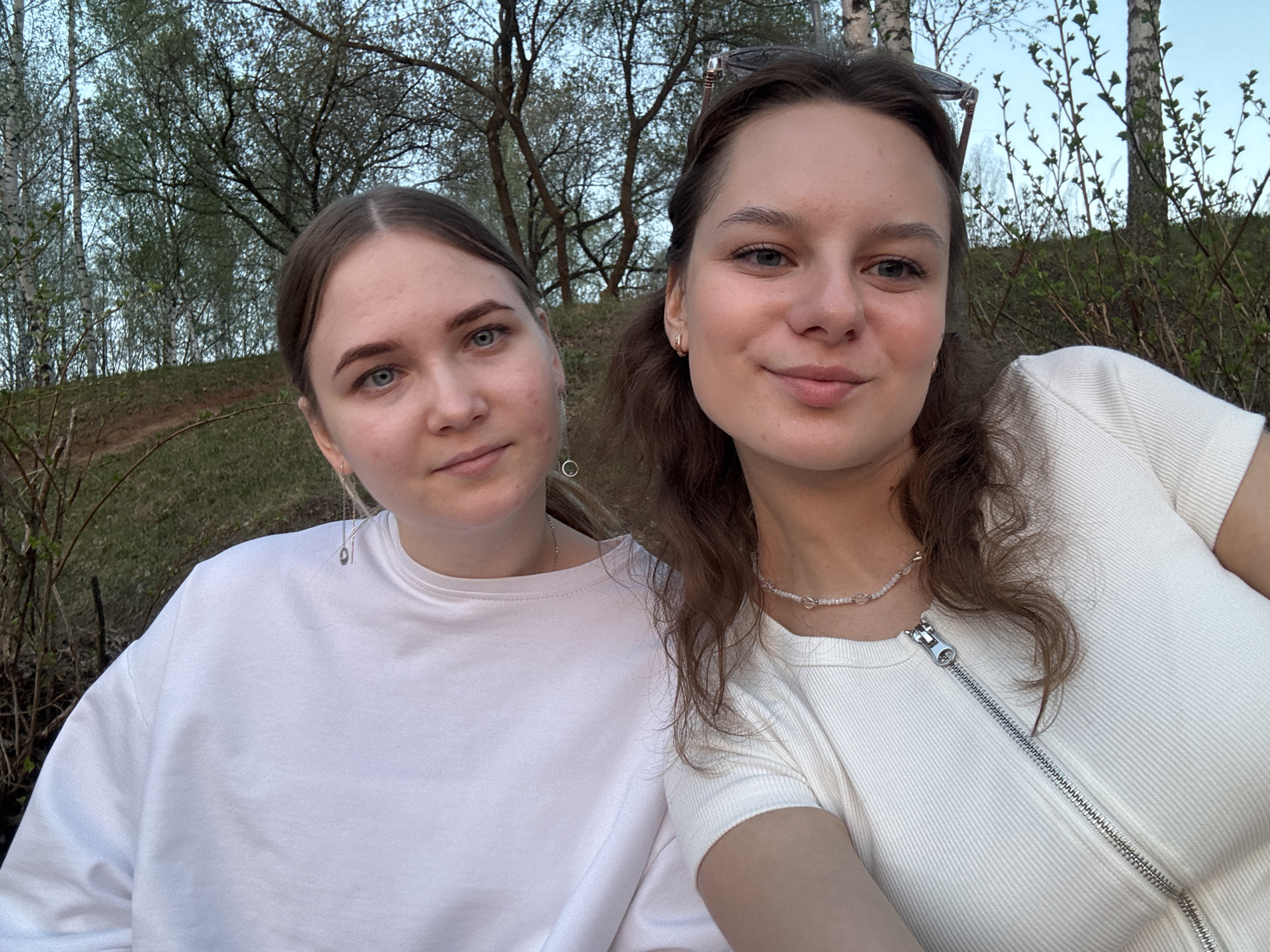 Two young women pose close together outdoors with trees and foliage visible in the background. The woman on the left has light brown hair pulled back, wearing a white sweatshirt and dangling earrings. The woman on the right has brown hair, some of which is pulled back with sunglasses perched on top of her head, and wears a white ribbed shirt with a prominent zipper down the front and a beaded necklace. The photo appears to be taken at a slightly upward angle. The location is likely in Chaikovsky, Russia based on the trees in the background.