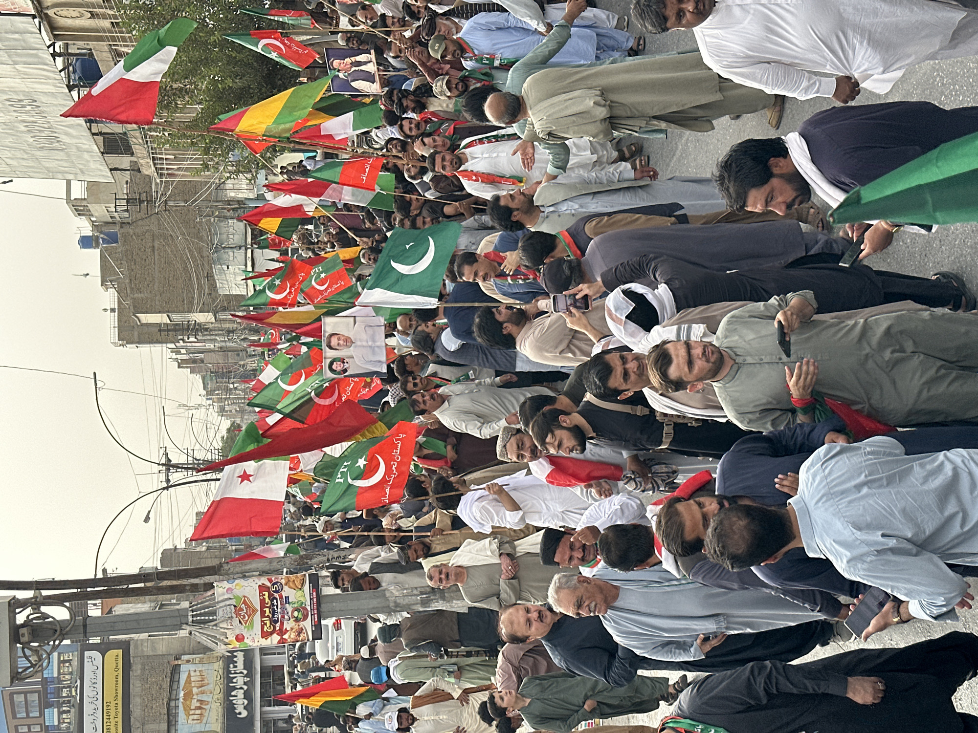 The image captures a street scene in Quetta City Tehsil, Pakistan, likely during a public gathering or procession. A dense crowd of men fills the street, many holding or standing near a multitude of flags. The flags are predominantly green, white, and red, with some featuring crescent moons and stars, indicative of Pakistan's national flag and political party affiliations. Several flags are also red and white, with a star emblem.
In the background, buildings line the street, suggesting an urban environment. A few vehicles are visible, including a white car and what appears to be a motorcycle. Overhead, a complex network of electrical wires crisscrosses the sky. Several posters are affixed to a utility pole in the foreground. One poster features an image of a man in traditional attire, along with text in what appears to be Urdu. Another poster displays images of food and beverages.
The people in the crowd are dressed in various traditional South Asian attire, including shalwar kameez. Many appear to be engaged in conversation or observing the surroundings. Some are holding mobile phones, either for communication or photography. The atmosphere seems active and potentially political, given the presence of numerous flags and the organized nature of the gathering. The lighting suggests it is daytime, with overcast conditions.
