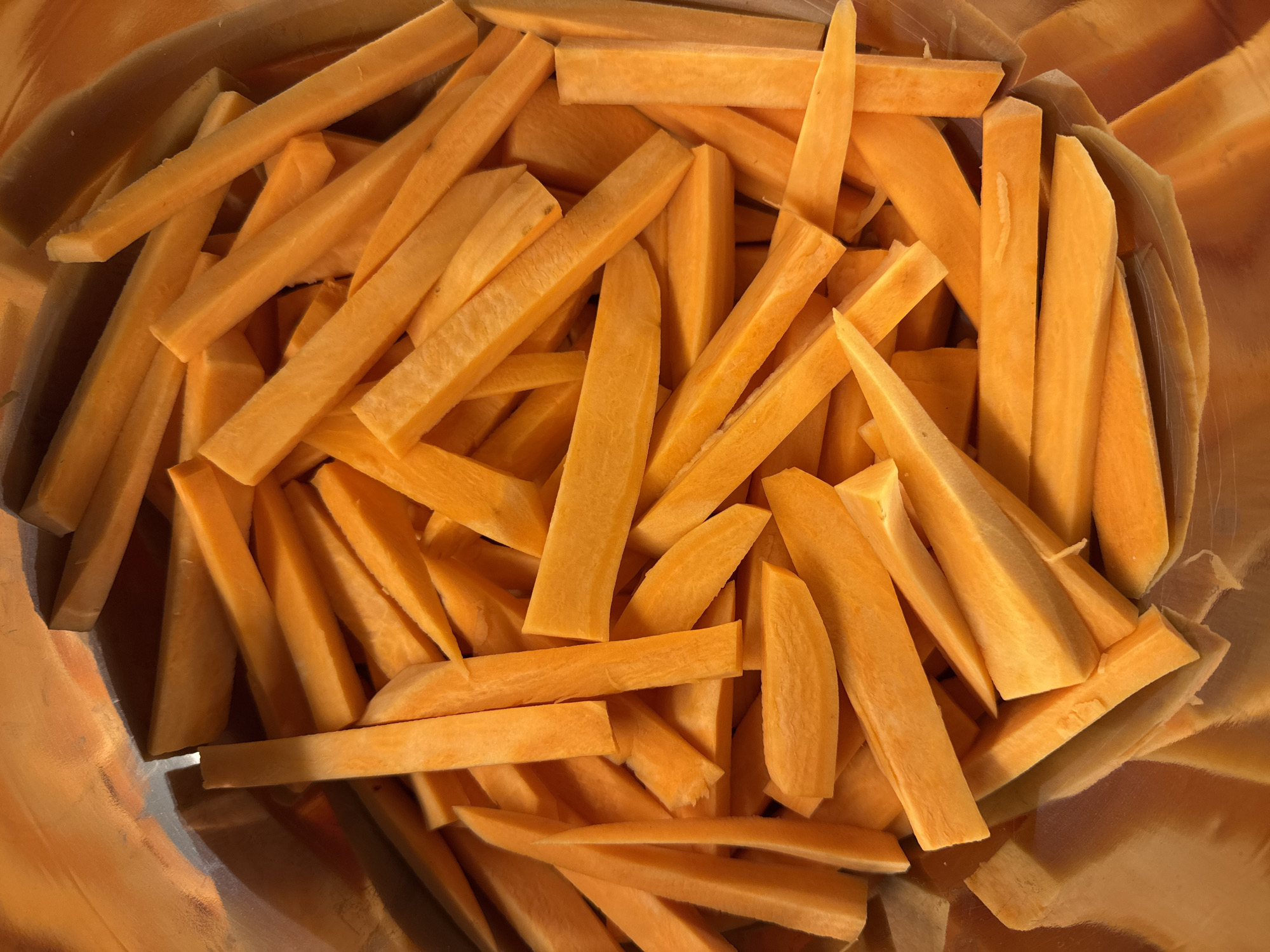 The image shows a close-up, overhead view of uncooked sweet potato fries filling a container. The fries are mostly orange in color and cut into stick shapes, varying in length and thickness. Some are blunt-ended while others have pointed tips. The container appears to be metallic or reflective, with light reflecting off its surface, creating an orange hue around the edges. The composition fills the frame, with no other objects or people visible. The location is not identifiable from the image.