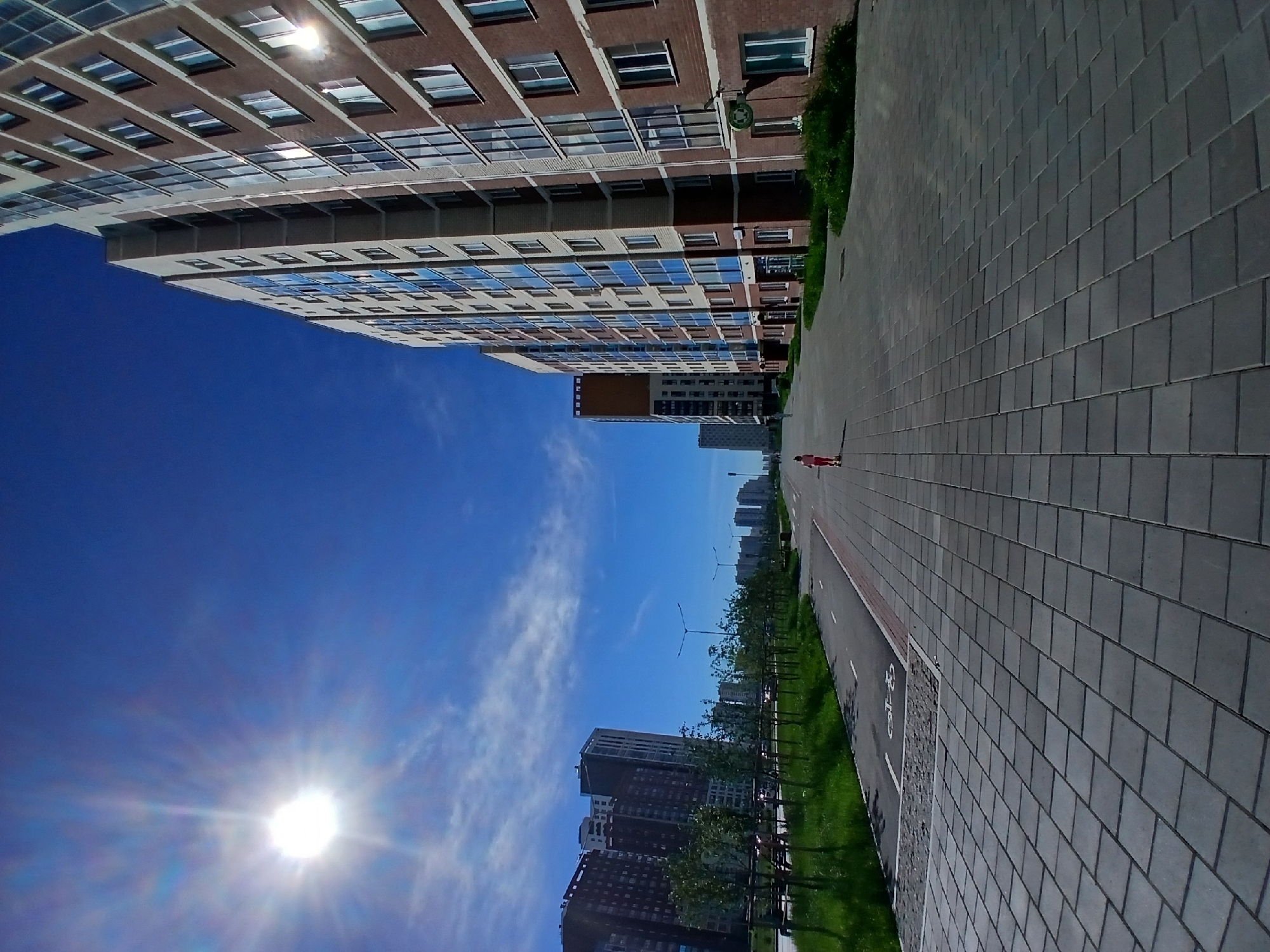 The image shows a bright, sunny day with a clear blue sky and a few wispy clouds. A prominent sunburst is visible in the upper left quadrant of the sky. In the foreground, a wide, grey paved walkway stretches into the distance, lined with dark grey rectangular pavers. To the left of the walkway is a strip of green grass with young trees planted in neat rows. Beyond the grass is a paved path with a bicycle symbol, and a road with some distant cars.
On the right side of the walkway, a large, multi-story brick building with numerous windows rises towards the sky. Its facade is a mix of brown brick and lighter-colored sections, with blue-tinted windows reflecting the sunlight. Further in the distance, other similar residential buildings are visible, creating a cityscape.
A small child, wearing a pink outfit, is riding a scooter on the walkway, moving away from the camera. The child is the only clearly discernible human subject. The scene appears to be a modern urban development, likely a residential area in Yekaterinburg, Russia, given the architectural style and the clear, sunny weather. The overall impression is one of a contemporary, planned urban environment on a pleasant day.