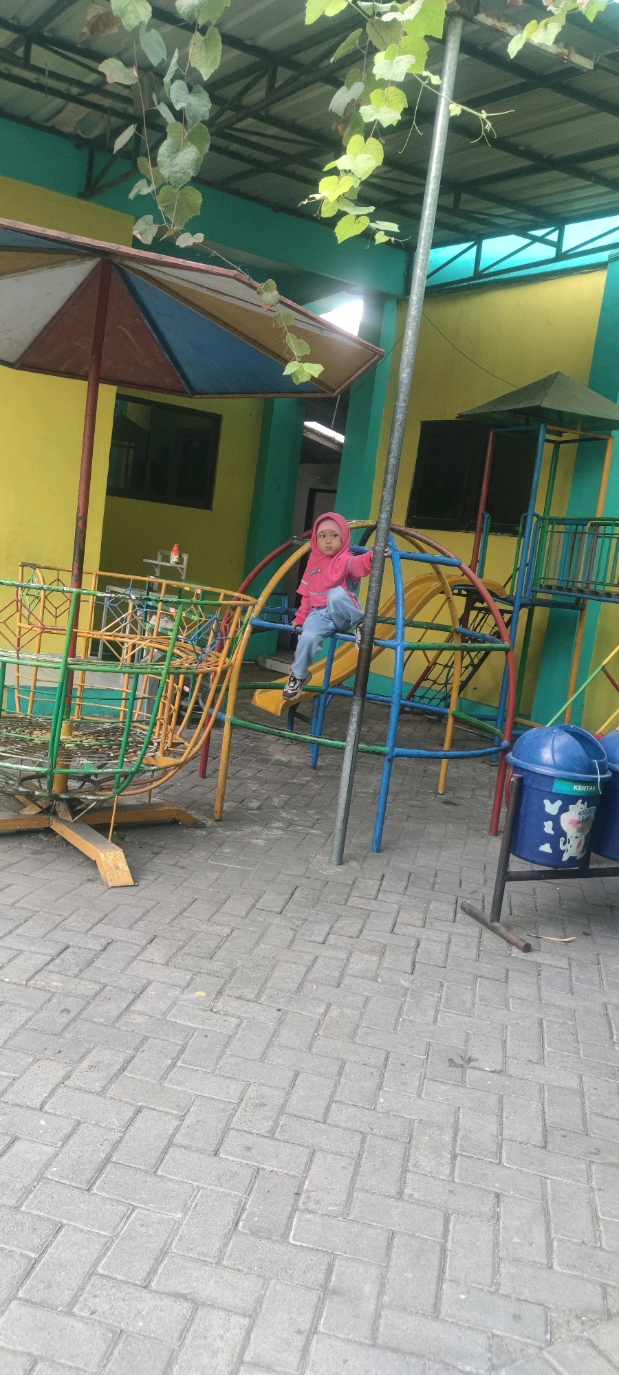 A young child, appearing to be of toddler age, is sitting on a blue metal bar of a colorful playground structure. The child is wearing a pink hooded jacket and light blue pants, with black and white sneakers. The playground equipment is made of metal with various colored bars, including yellow, blue, and red, and appears to be situated outdoors in a paved area with interlocking brick pavers. Behind the playground, a yellow building with teal trim is visible, featuring windows with dark panes. Overhanging the scene are green leafy vines and a section of a metal roof structure. A blue trash can with a white cow graphic and the word "KERTAS" (meaning paper) is visible to the right of the playground. The setting is likely a public space or an outdoor area associated with a school or community center in Banjarsari, Indonesia.