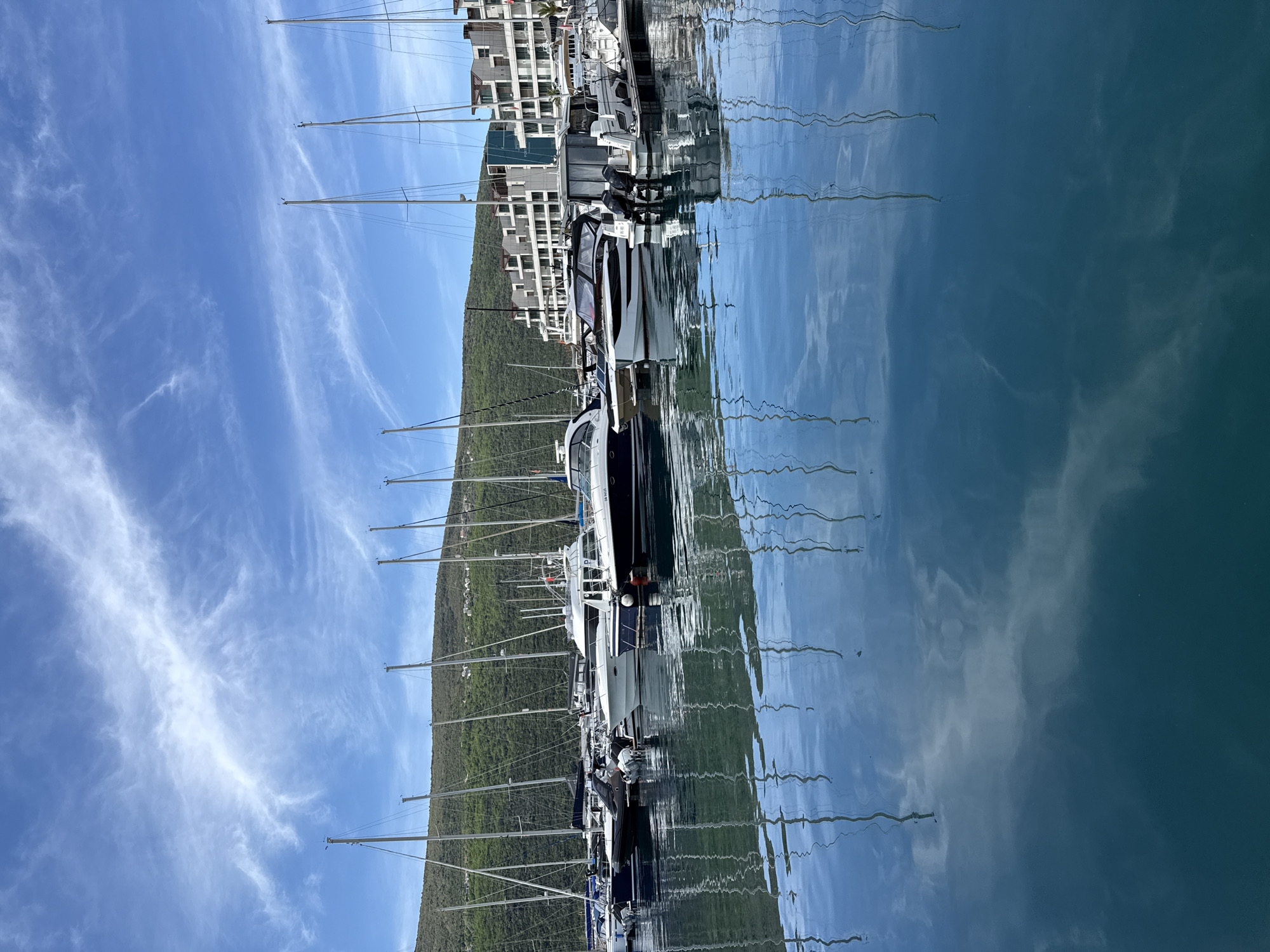 The image depicts a marina in Đenovići, Montenegro. Several yachts and sailboats are docked in the water. The reflections of the boats and masts are visible on the water's surface. A building stands on the edge of the marina, with a green hillside behind. The sky is blue with scattered clouds.